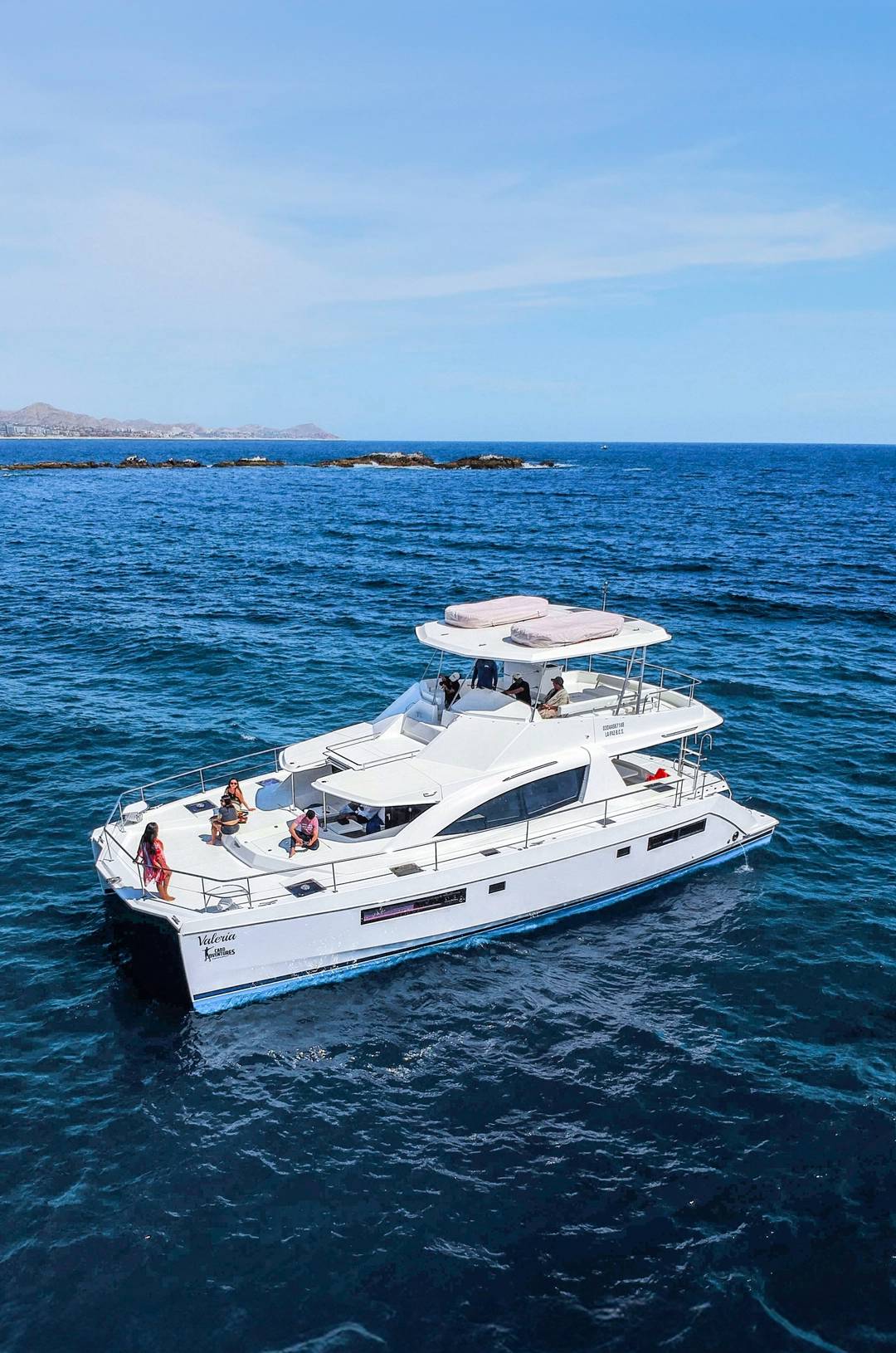 A luxury yacht cruises the blue waters near Cabo, with people relaxing on deck under a clear sky.