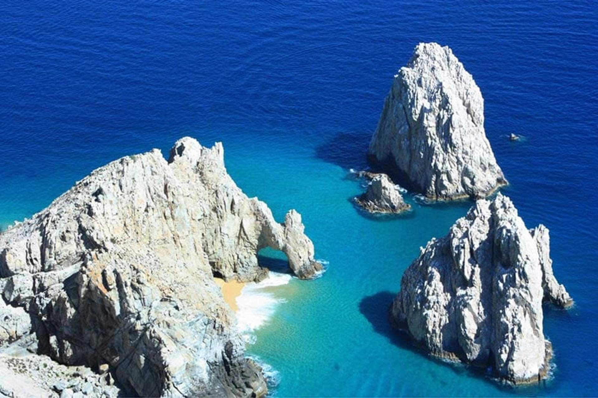 An aerial view of the iconic rock formations at Land's End in Cabo San Lucas, Mexico. The archway in one of the rocks frames a secluded beach with golden sand and turquoise water.