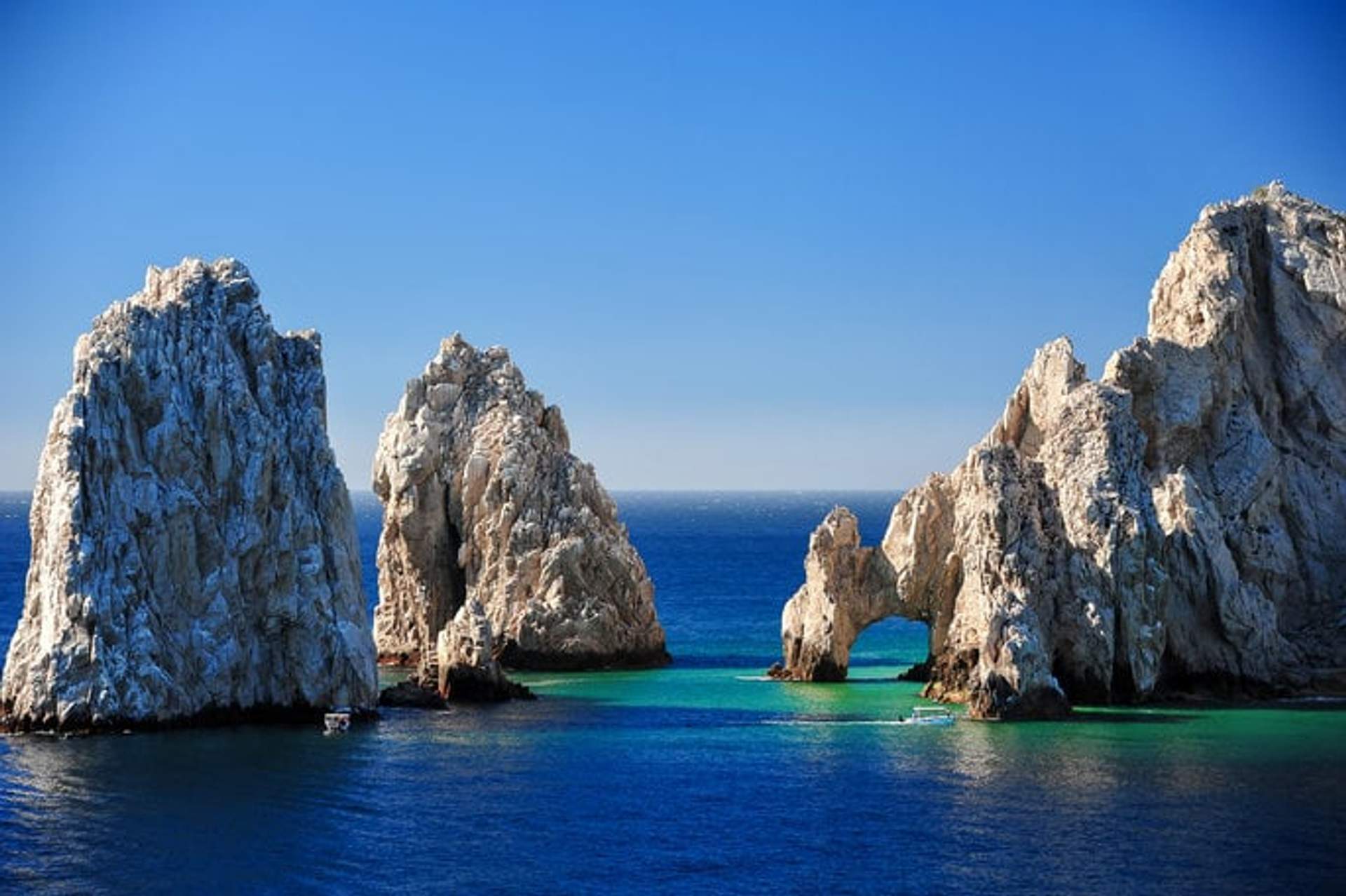 A scenic view of El Arco de Cabo San Lucas in Mexico, showcasing towering rock formations rising from the clear blue waters of the Pacific Ocean under a bright, clear sky. The famous arch is prominently featured, with vibrant turquoise waters surrounding the natural landmark.