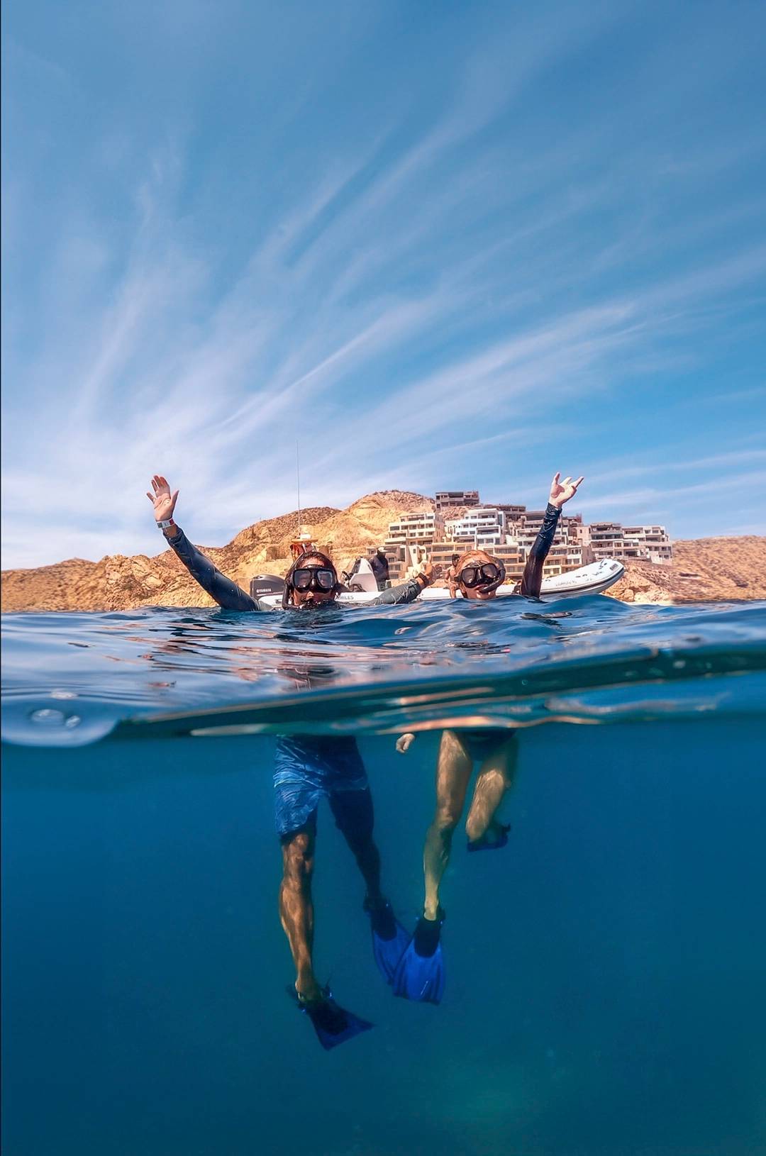 Two snorkelers with raised arms, partially submerged in the water, with a rocky coastline and buildings in the background.