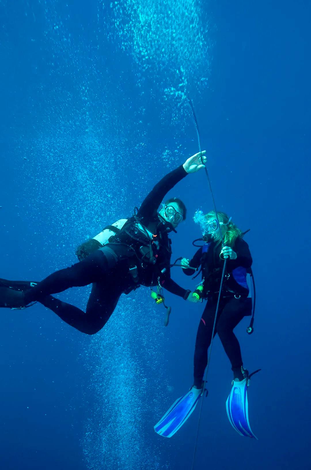 Two scuba divers in full gear hold onto a guide rope underwater, surrounded by bubbles, in deep blue ocean water.