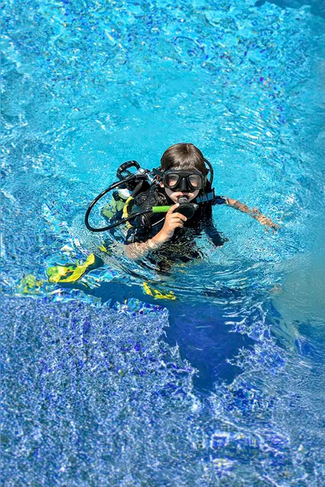 Young diver practicing with scuba gear in a swimming pool, surrounded by clear blue water.