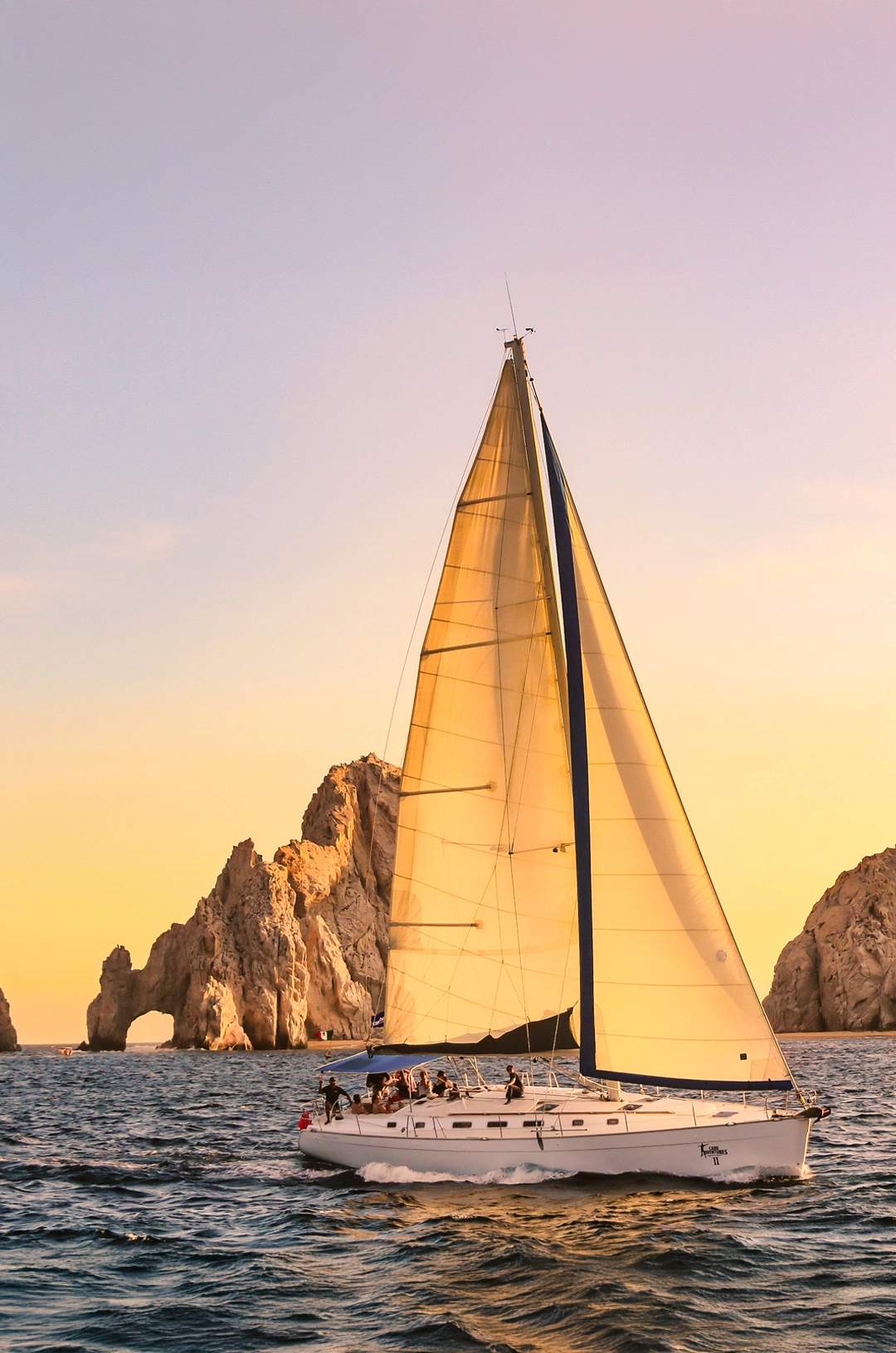 Sailboat cruising near Cabo's Arch at sunset with golden skies and rocky formations in the background.