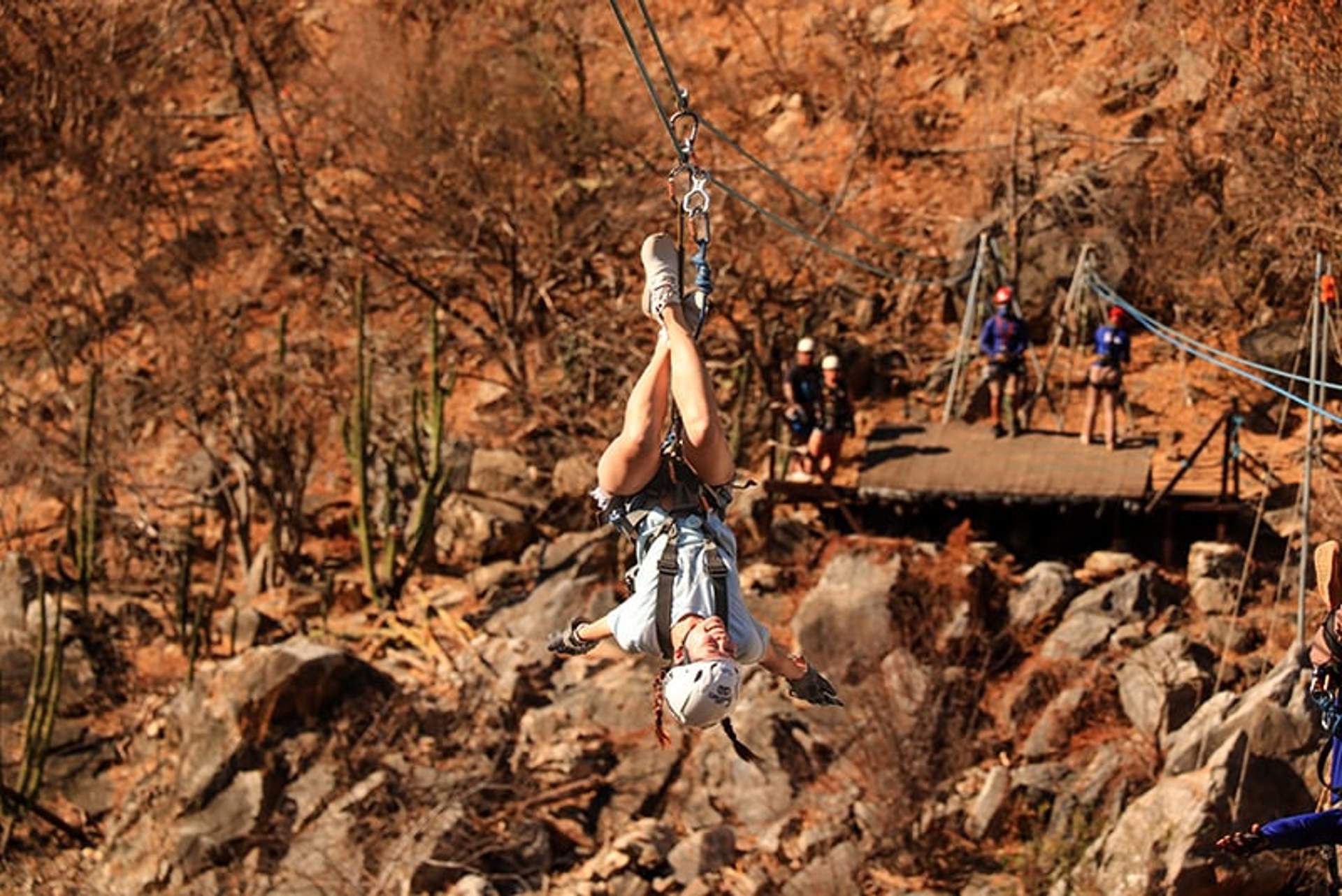 A person zip lines through a rugged, rocky terrain, hanging upside down for an adventurous thrill. They are wearing a helmet and safety gear, with other participants and the zip line platform visible in the background amidst a dry, desert-like landscape.