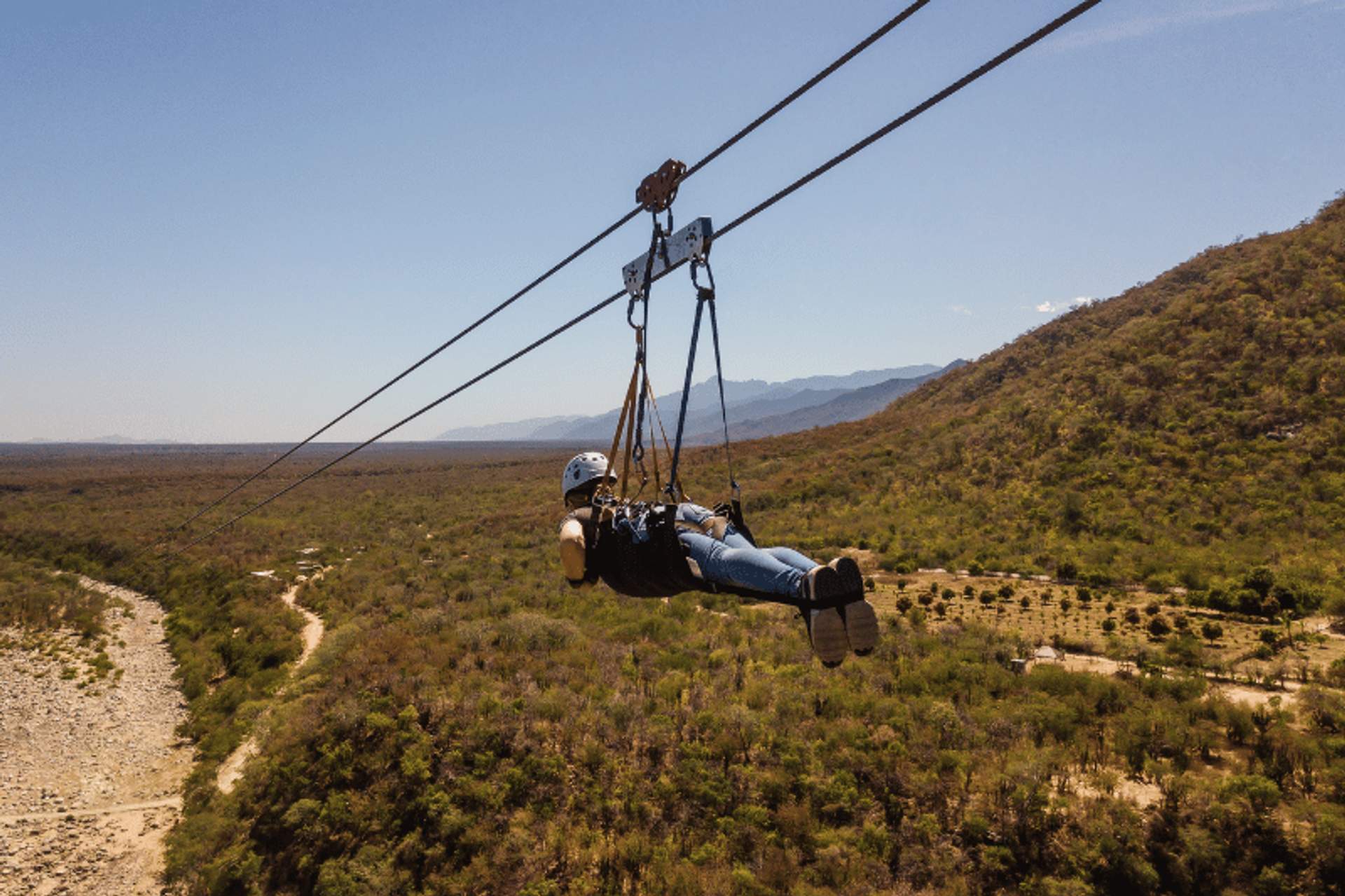 Person zip-lining over a scenic landscape with hills, trees, and a dry riverbed, enjoying an adventure activity in Cabo San Lucas.