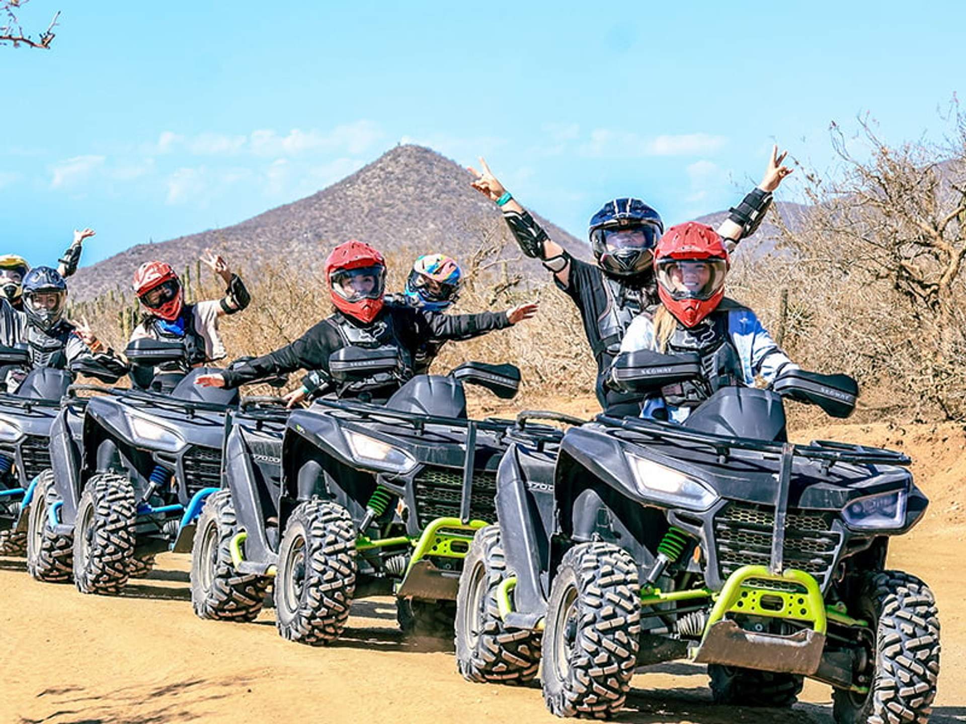 A group of people wearing helmets and protective gear ride all-terrain vehicles (ATVs) on a dirt trail with a mountain in the background.