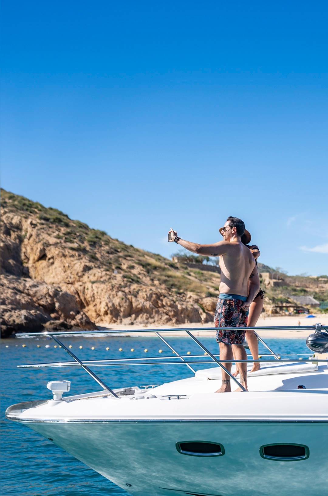 A couple on a yacht takes a selfie with a rocky coastline in the background under a clear blue sky, enjoying the sunny day.
