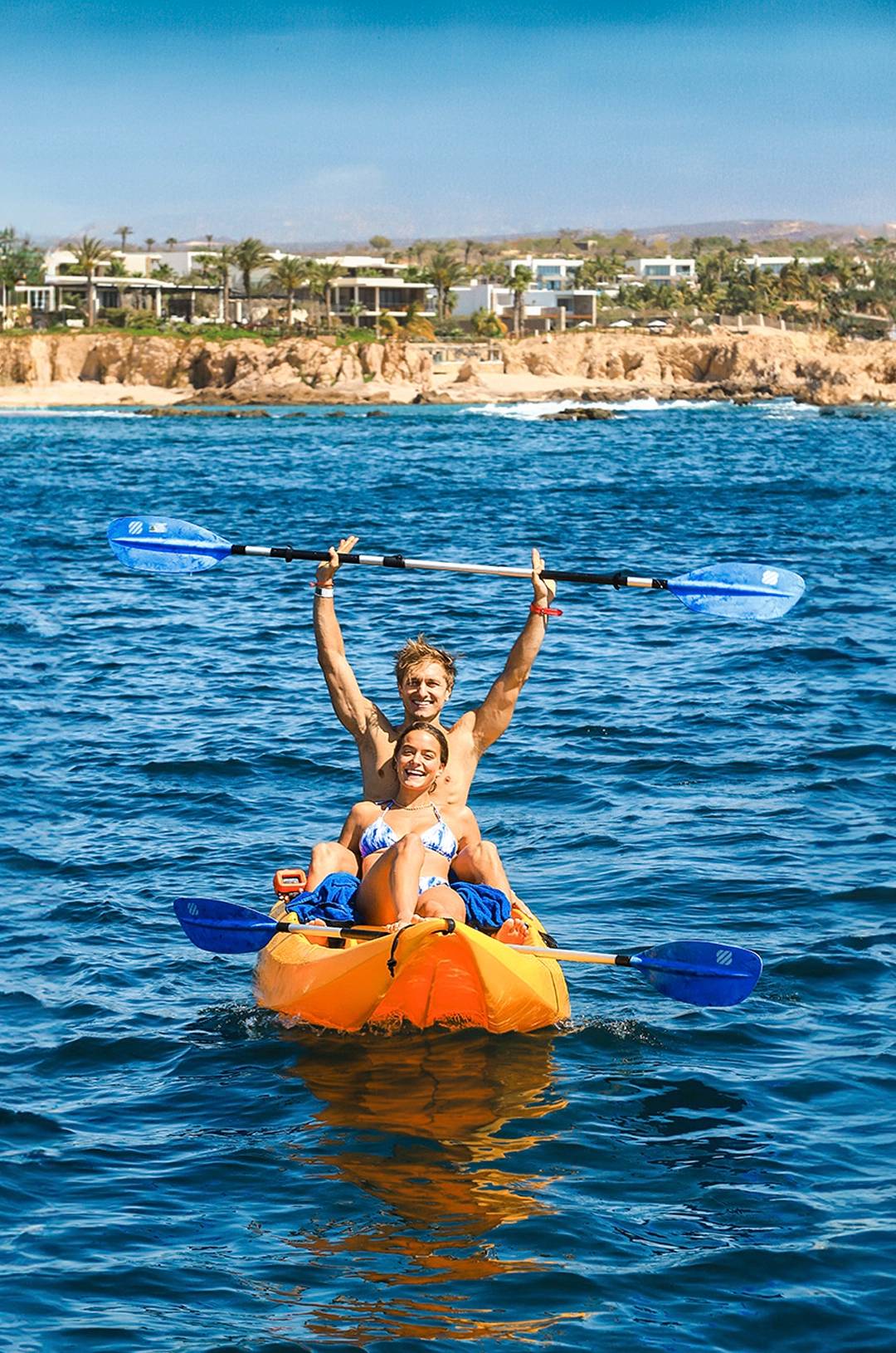 A couple kayaking on the ocean, smiling and holding their paddles up, with a rocky coastline and houses in the background.