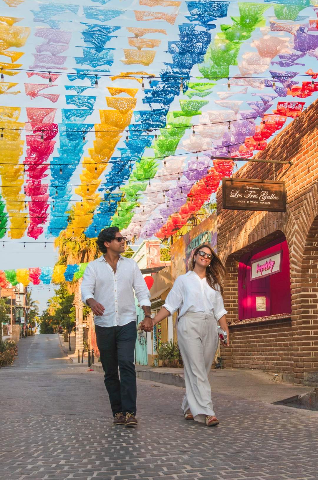 A couple walks under colorful papel picado banners on a cobblestone street beside shops and a brick building.