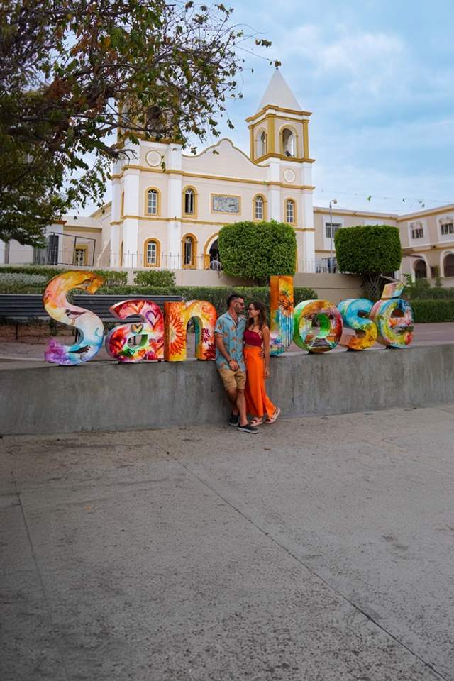 Tourists pose by a colorful "San José" sign in front of a historic church in Los Cabos.