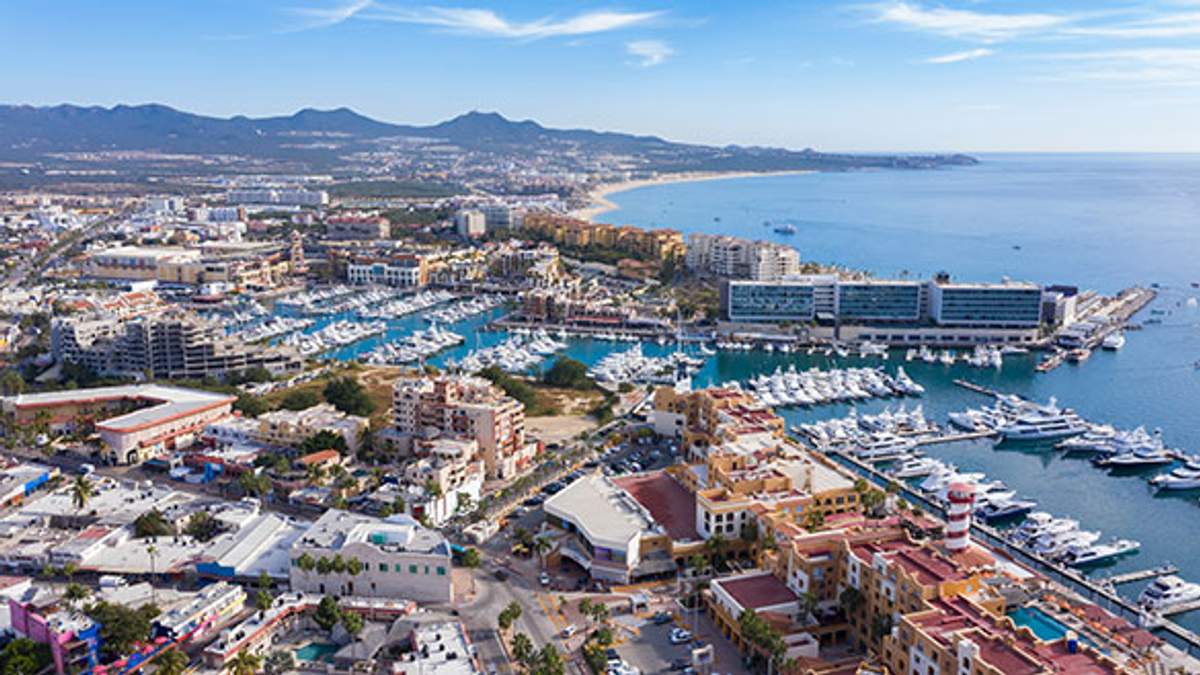 A panoramic aerial view of Cabo San Lucas marina, showcasing a bustling harbor filled with boats and yachts, surrounded by vibrant buildings and set against a backdrop of mountains and a curving coastline.
