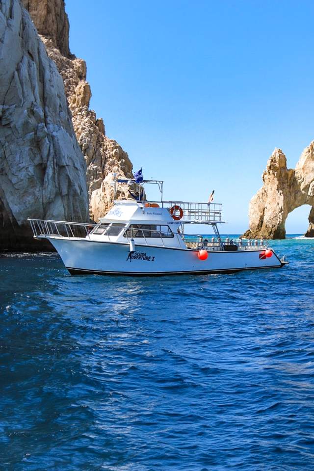 A white boat labeled "Scuba Adventure" floats near rugged cliffs and the iconic rock arch at Cabo San Lucas under a blue sky.