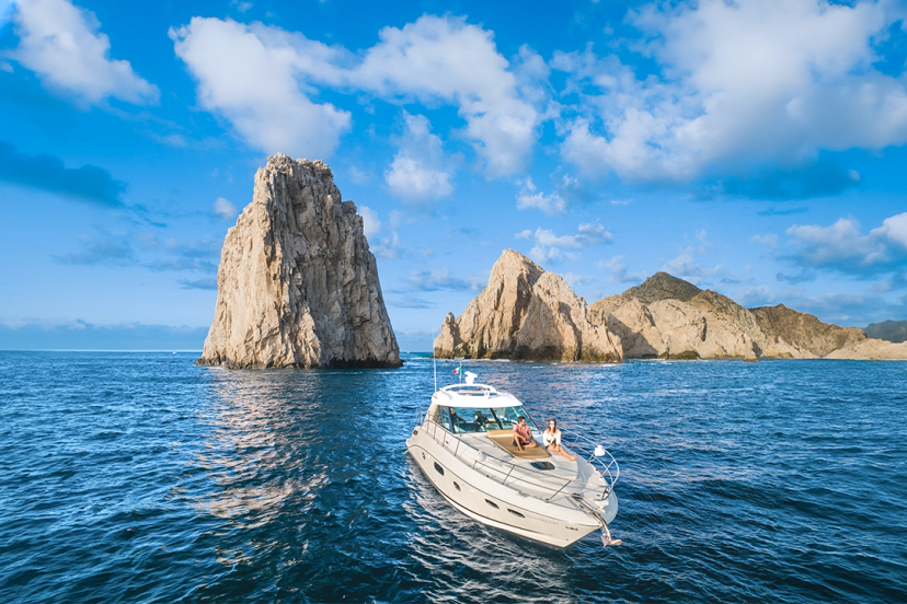 Boat Cruising In Cabo's Clear Blue Waters With Stunning Rock Formations In The Background Under A Bright Sky