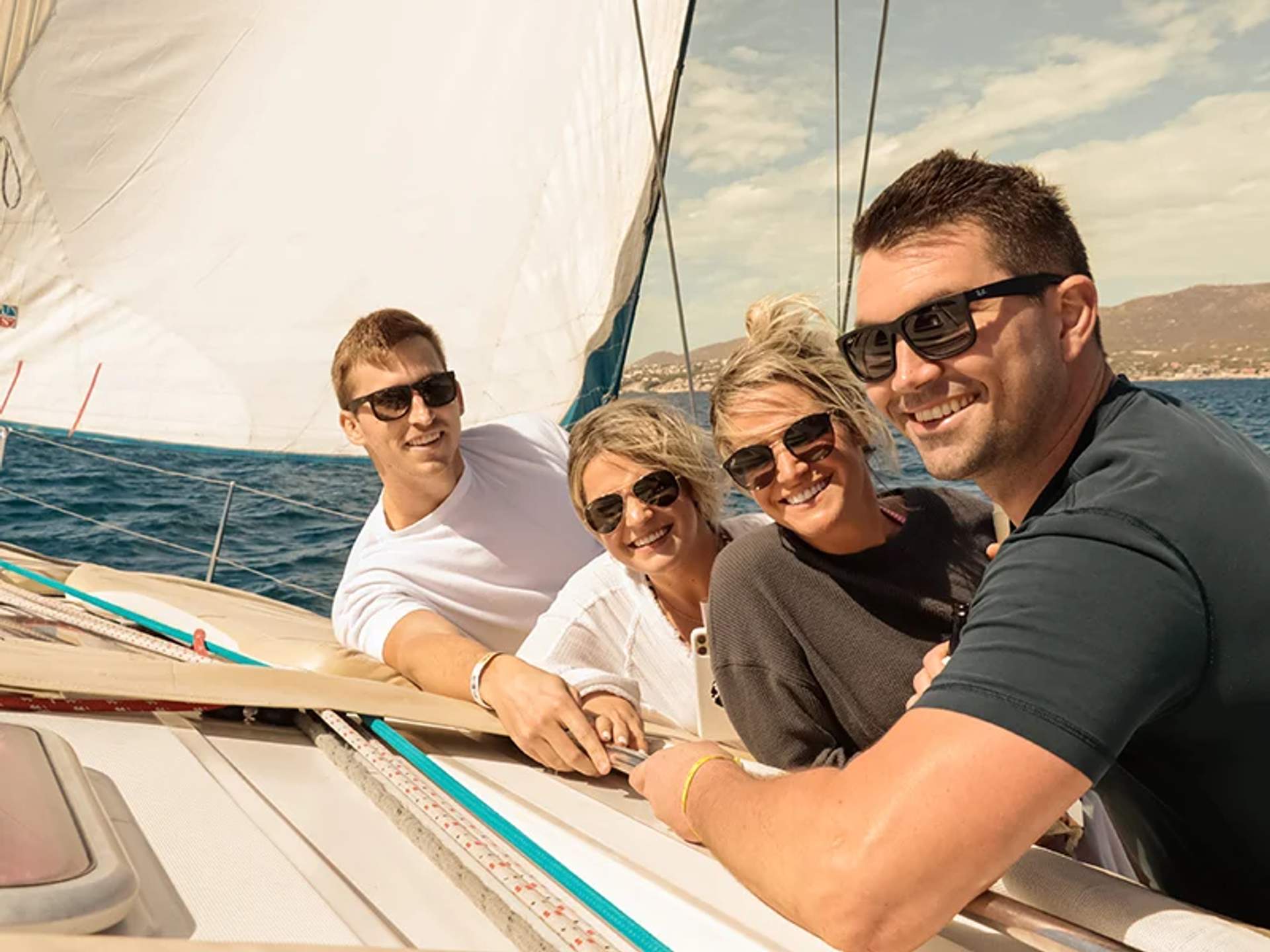A group of friends smiles for a photo while sailing on a sunny day, with the sail and ocean in the background.