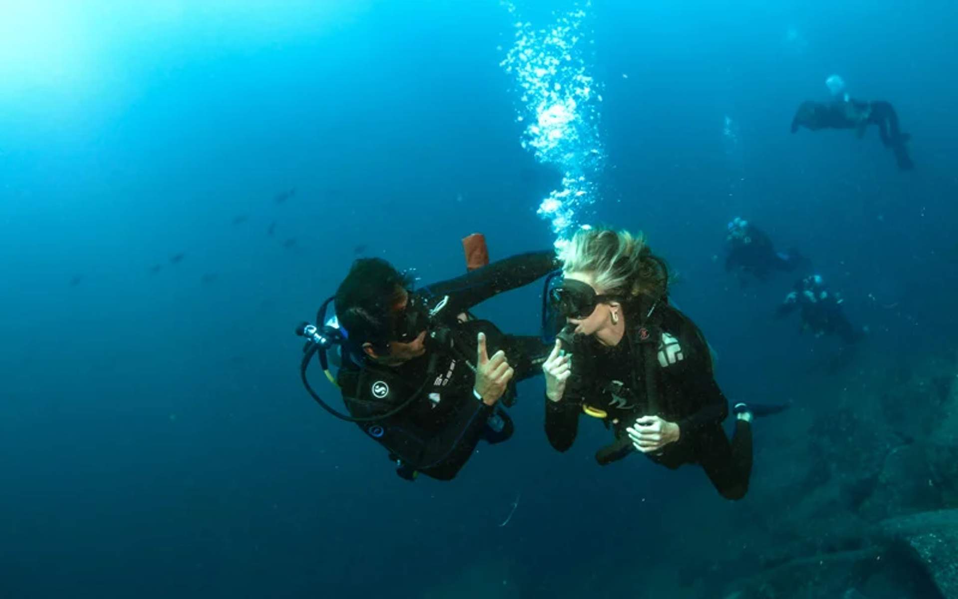 Two scuba divers underwater are communicating using hand signals, with bubbles rising above them and other divers visible in the background.