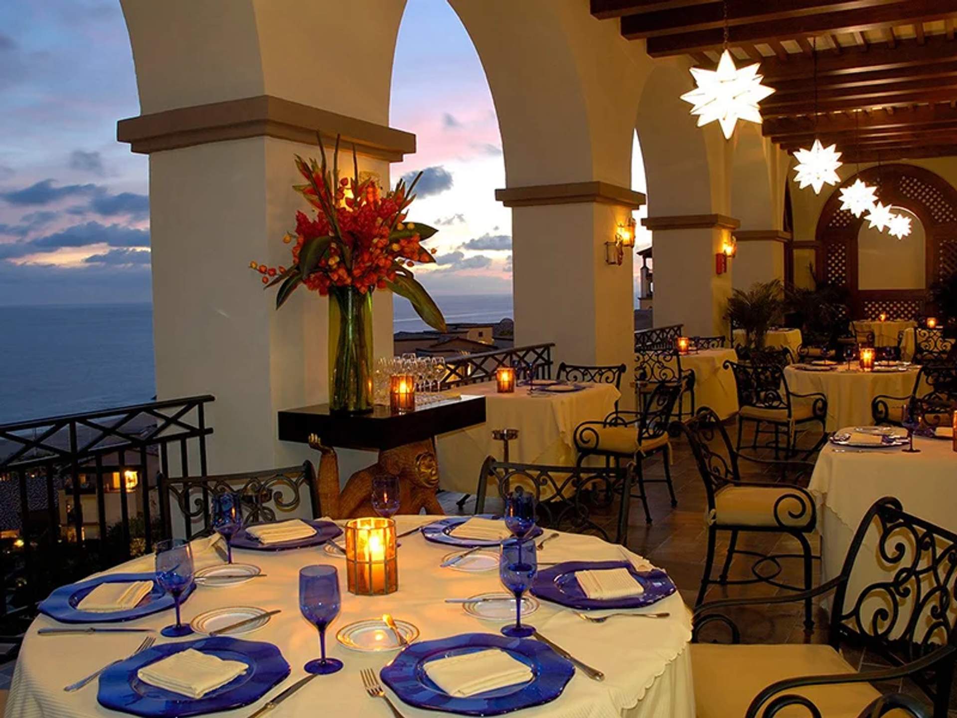 Elegant outdoor dining area at sunset, with candles and ocean view, set with blue glassware and white tablecloths.