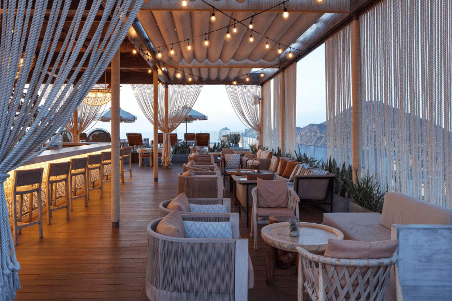 Elegant outdoor lounge area with wicker furniture and string lights, offering a cozy atmosphere with views of the ocean and mountains in Cabo San Lucas.