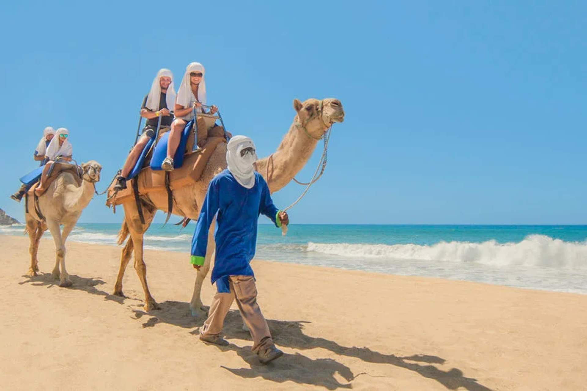 Tourists, wearing protective head coverings, are riding camels along a sandy beach, led by a guide dressed in blue. The scene showcases a sunny day with a clear blue sky and the ocean waves gently hitting the shore in the background.