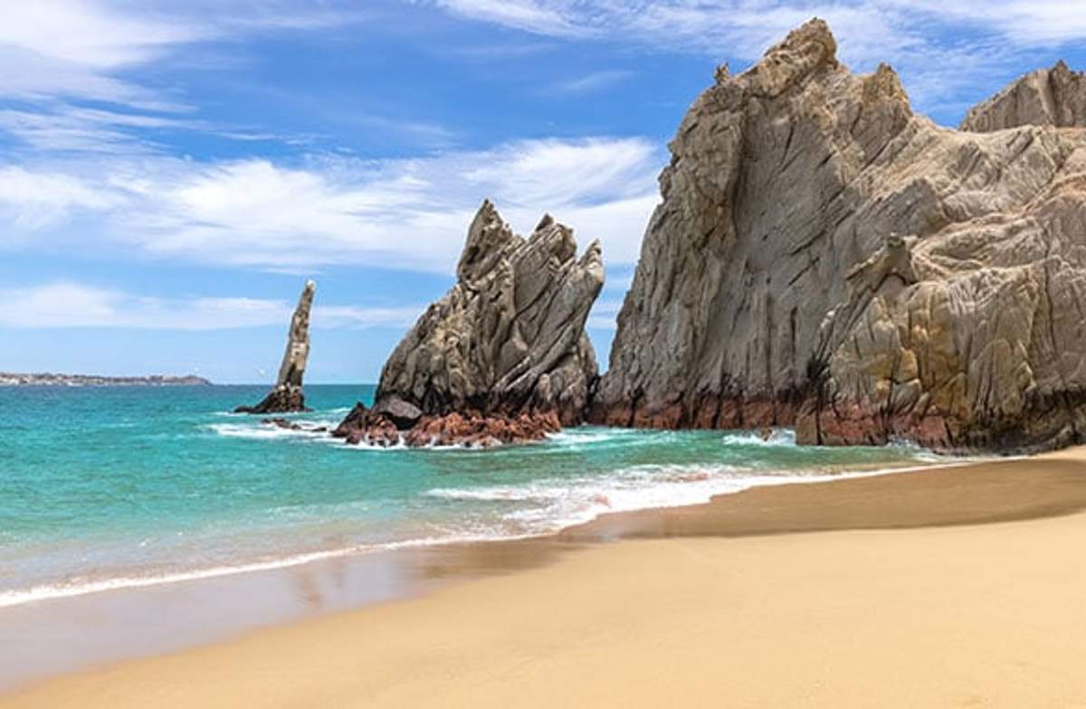 A pristine beach in Cabo San Lucas, featuring clear turquoise waters and dramatic rock formations jutting out of the sea. The golden sand stretches out under a bright blue sky with scattered clouds, creating a picturesque and inviting coastal scene.