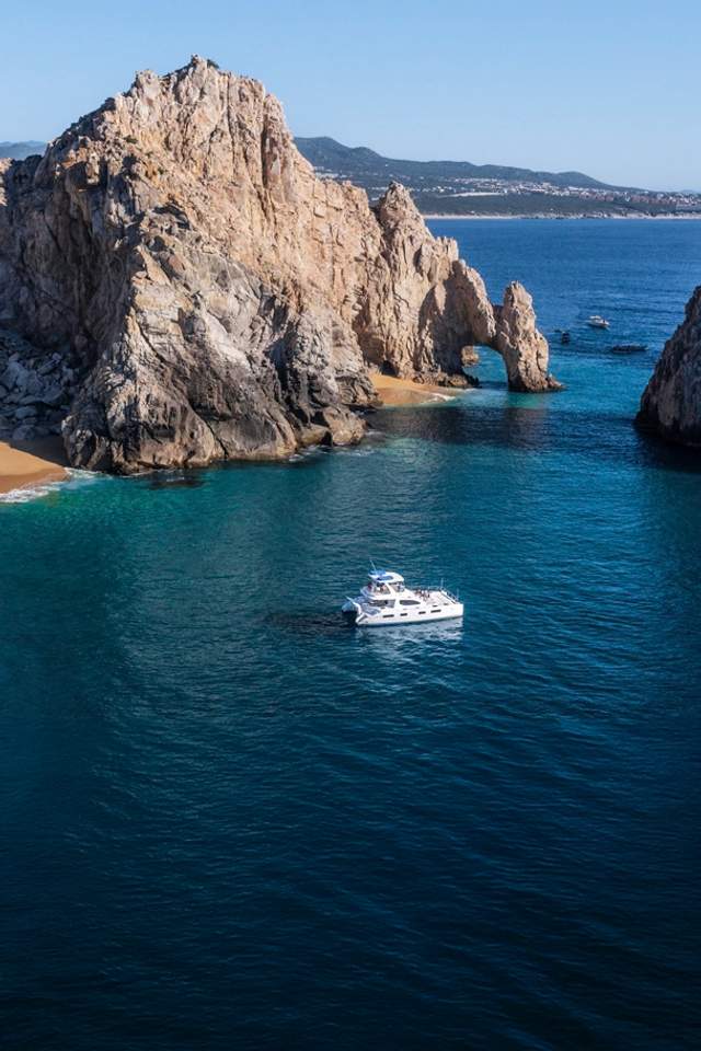 A boat sailing near the dramatic rock formations of Cabo San Lucas with clear blue water and mountains in the background.