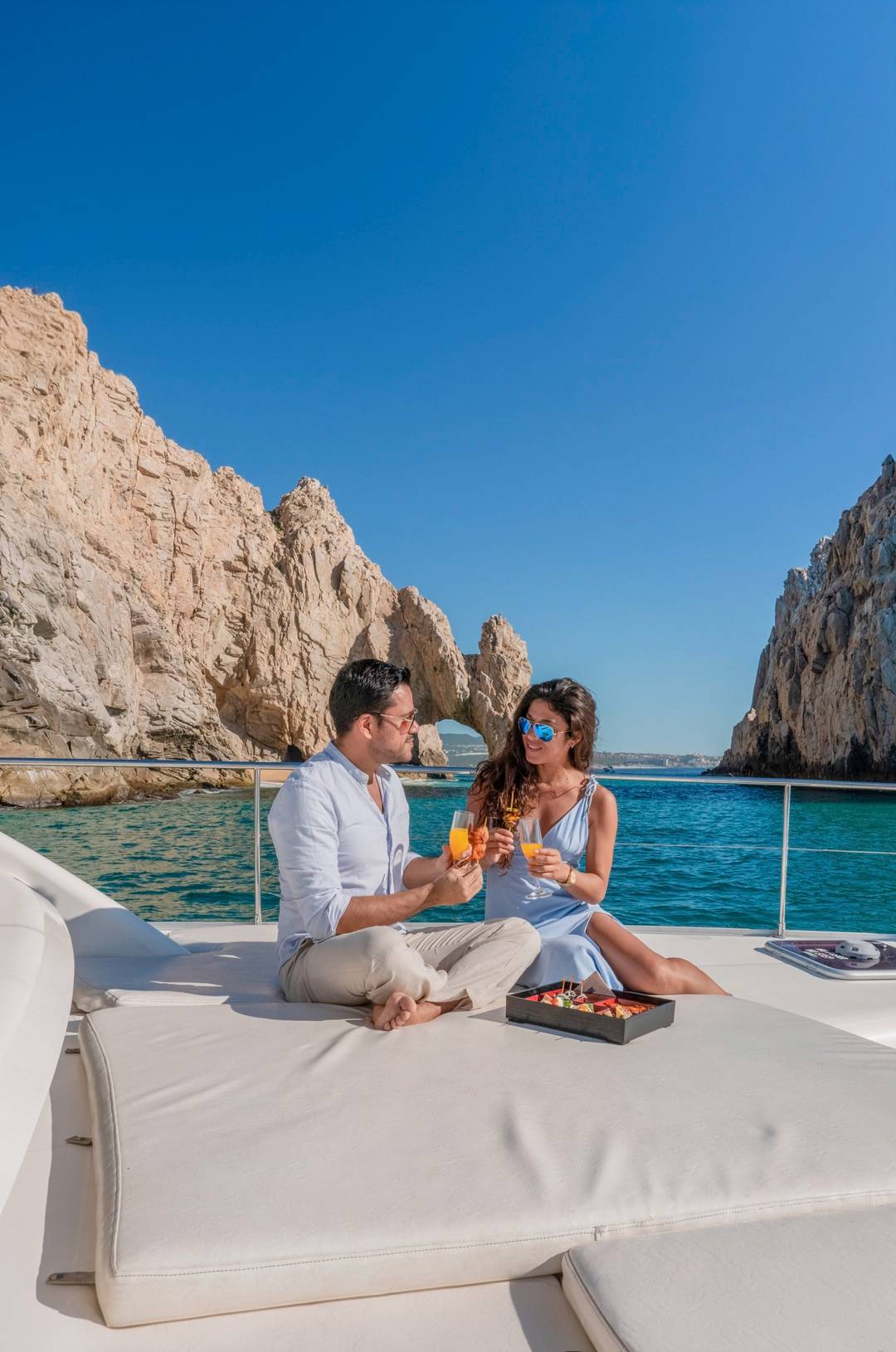 A couple enjoys drinks and snacks on a boat with a scenic view of rocky cliffs and blue sky.