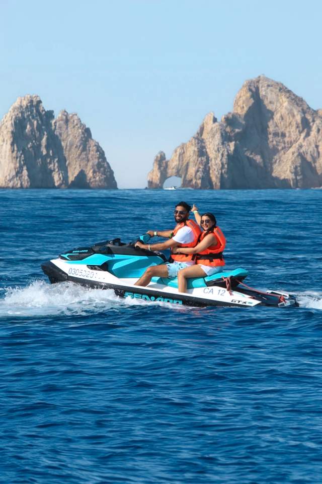 A couple rides a jet ski in the ocean with large rock formations in the background, both wearing orange life jackets.