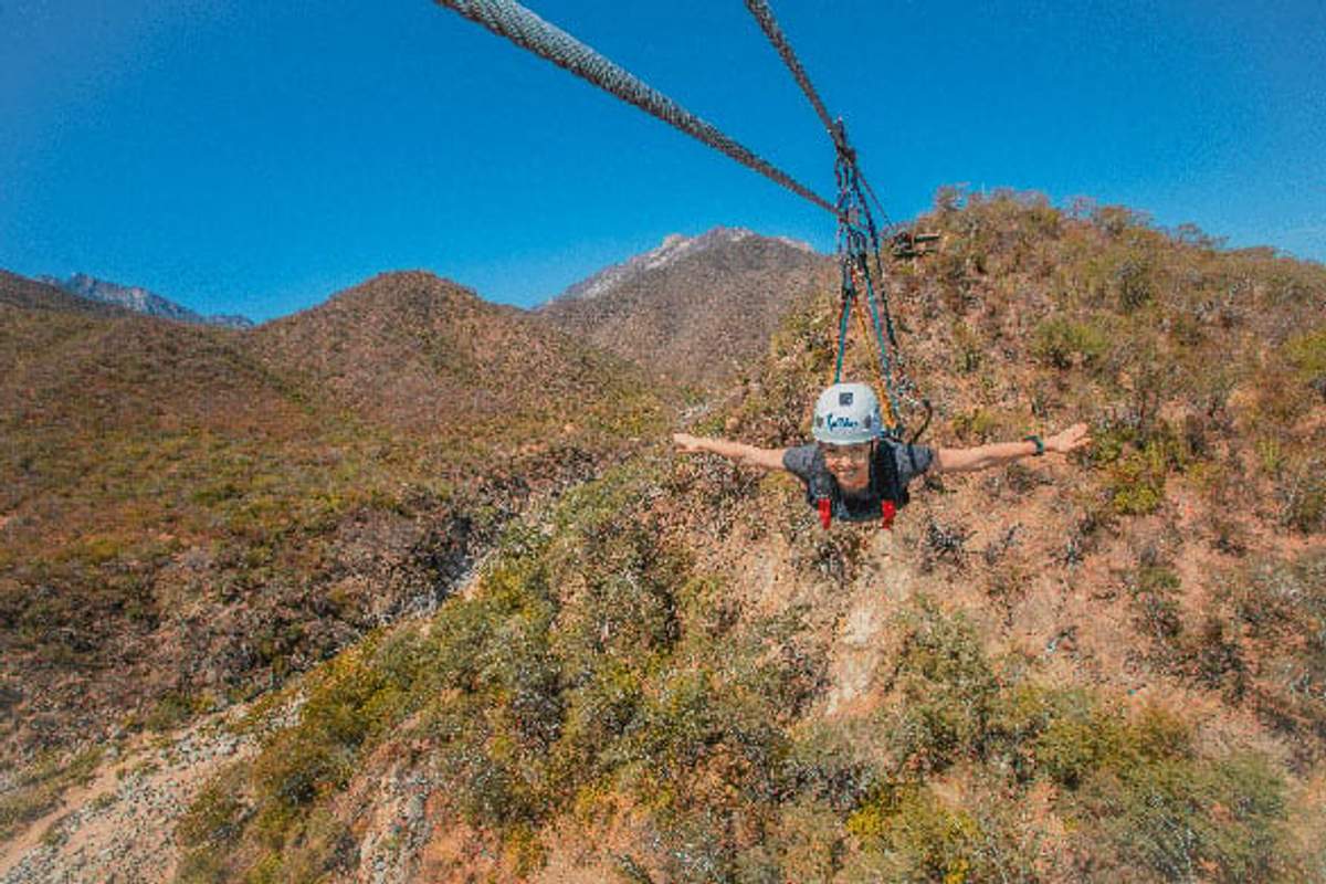 A person in a helmet and harness enjoys a thrilling zipline ride over a dry, mountainous landscape with scattered vegetation. The clear blue sky and distant hills enhance the adventurous setting.