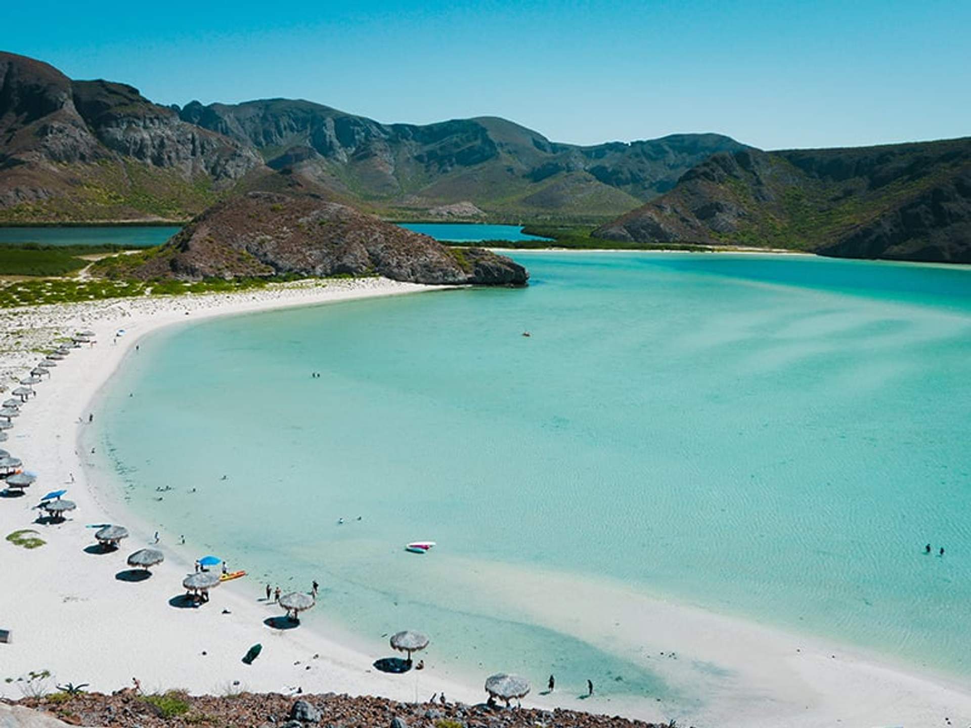 Aerial view of Balandra Beach with turquoise water, white sand, beach umbrellas, and surrounding mountains.