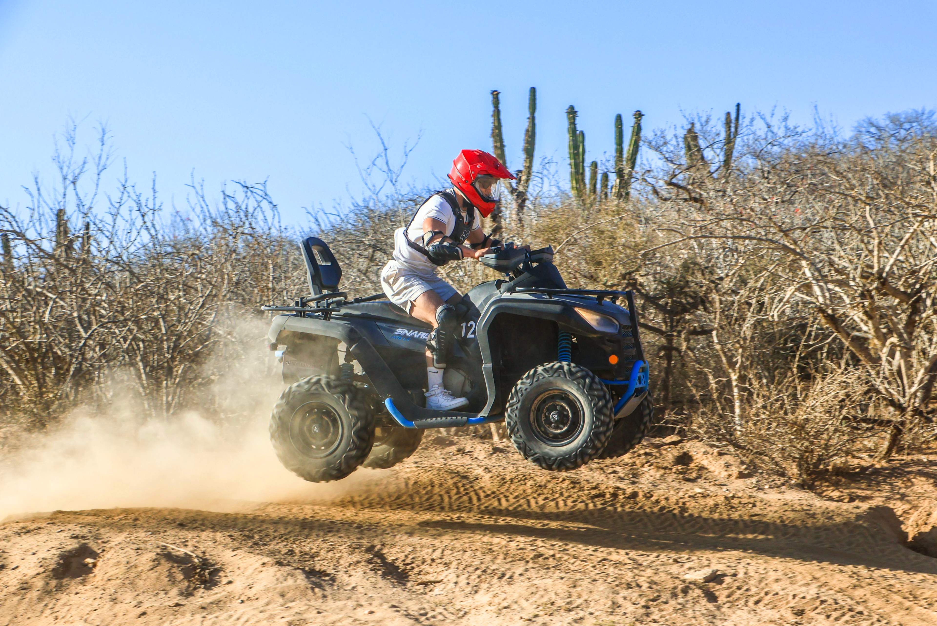 a person is seen riding an ATV while performing a jump on a sandy and arid terrain, surrounded by desert vegetation and cacti.