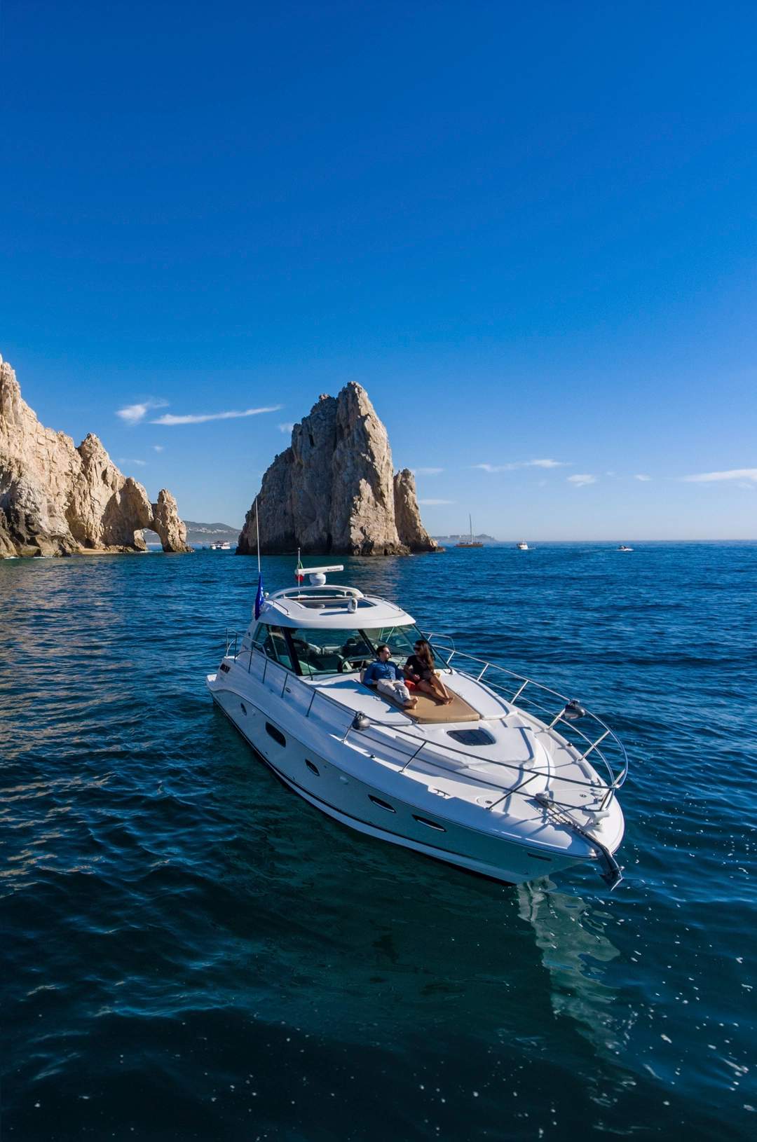 A luxury yacht cruises by the rock formations of Los Cabos under a clear sky, with two people relaxing on the deck.