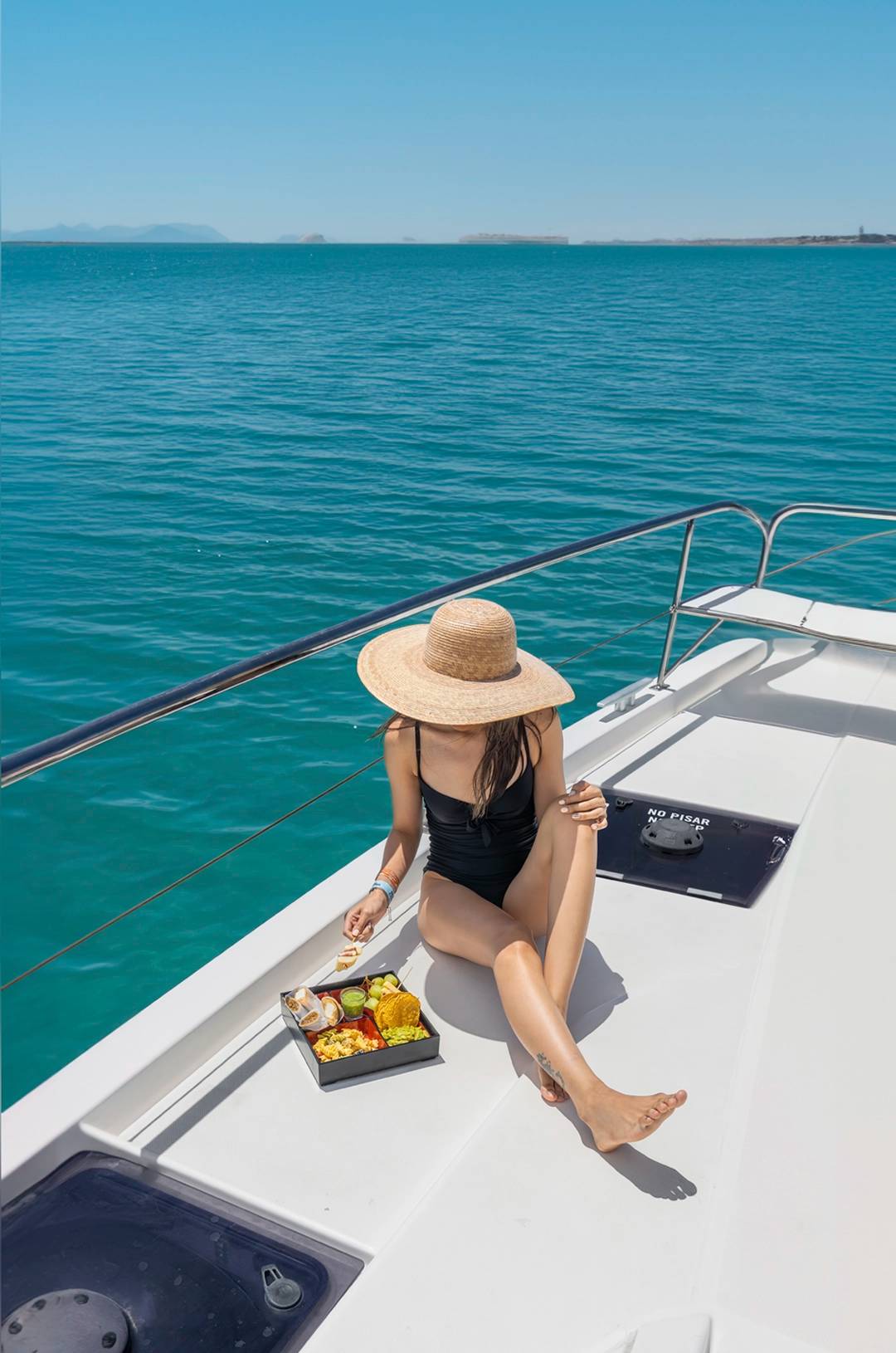 A woman in a black swimsuit and wide-brimmed hat enjoying a meal on the deck of a boat with blue waters in the background.