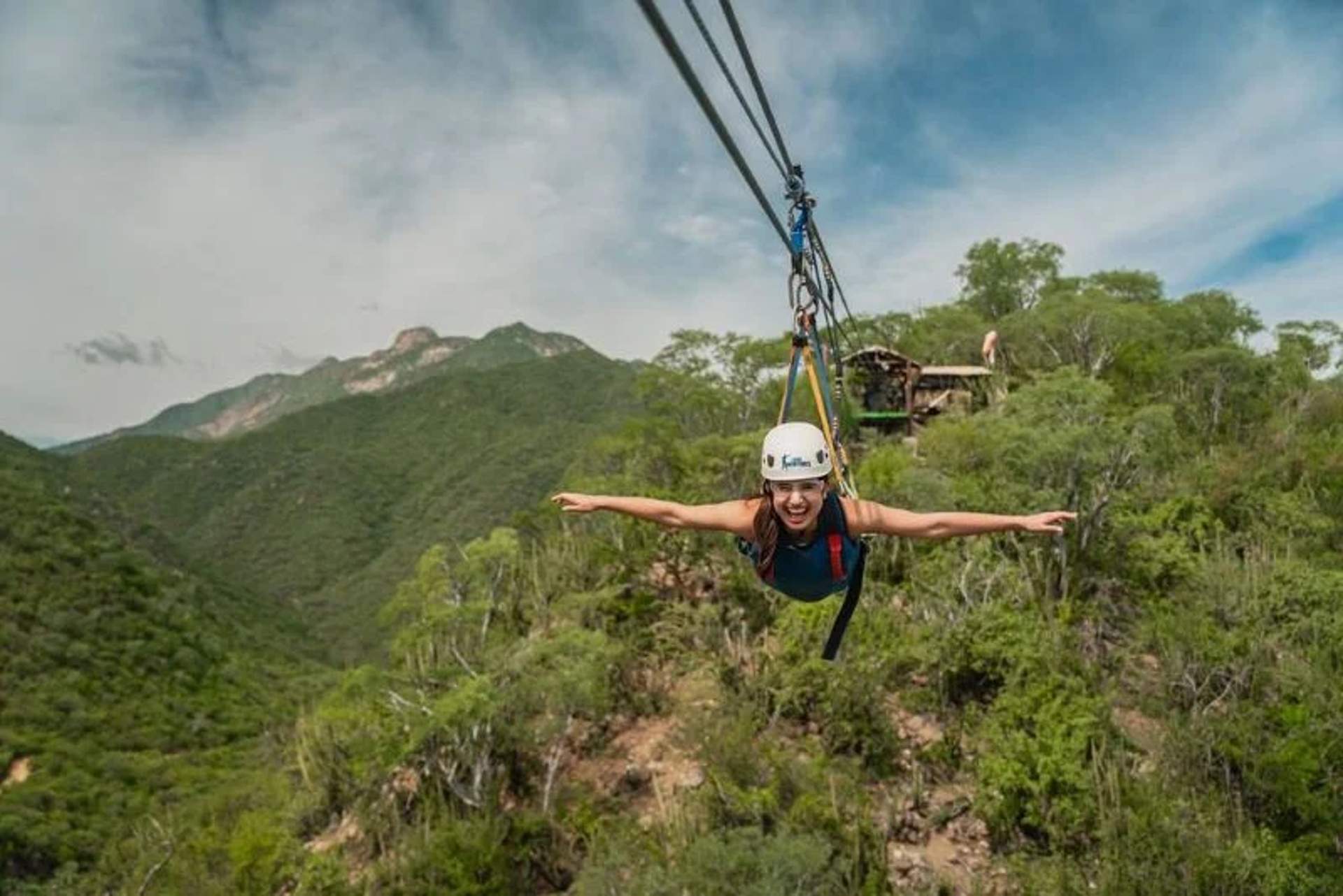 Person zip-lining over a lush green forest with mountains in the background, arms outstretched and smiling.