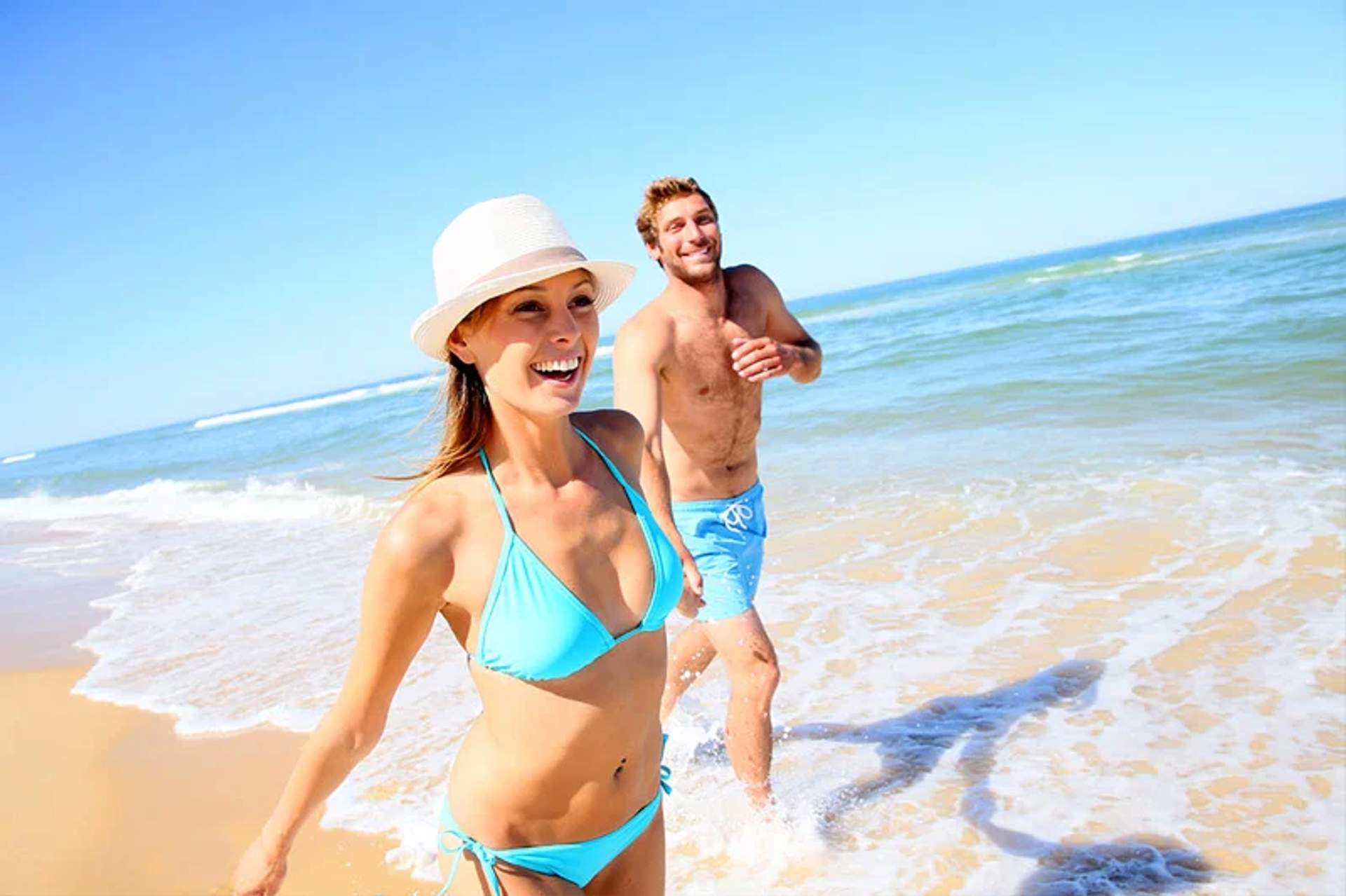 Young couple running and laughing along the shore of a sunny beach. She is wearing a light blue bikini and a white hat, while he is in light blue swim trunks. The sea and clear blue sky stretch out in the background.