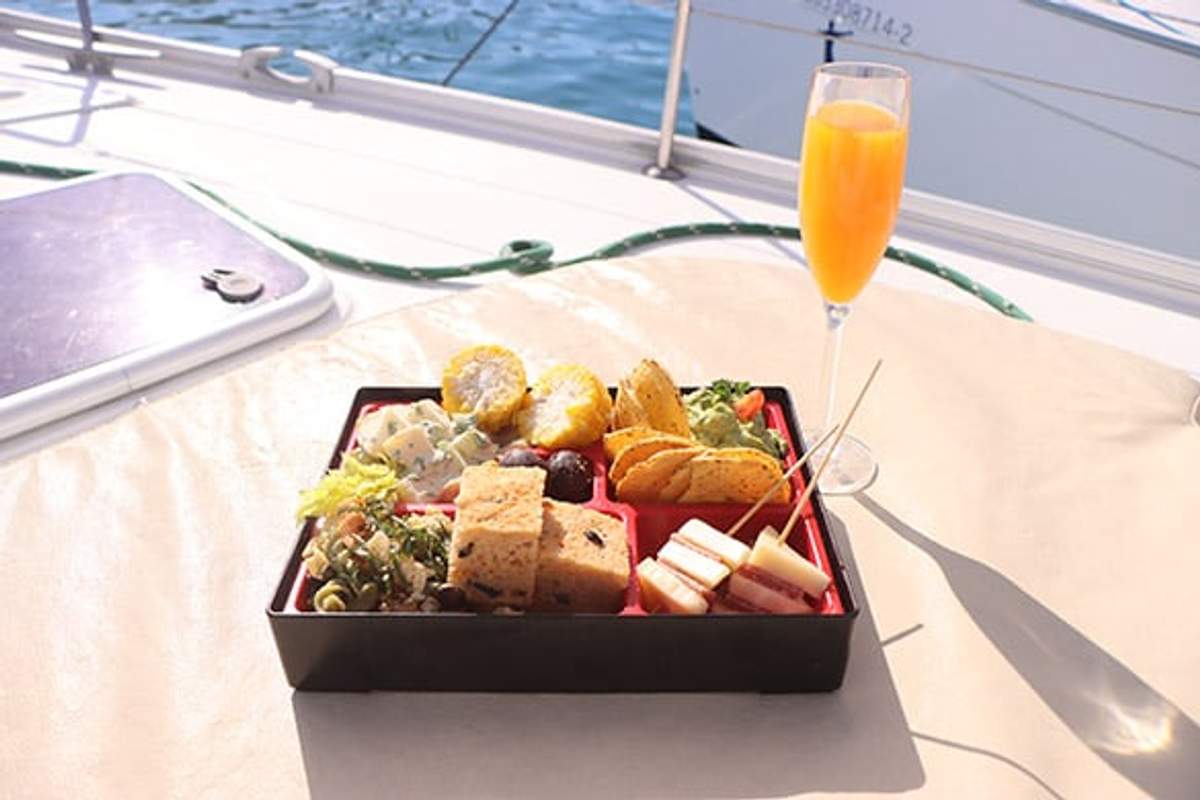 A bento box with assorted foods and a glass of orange juice on a yacht, with the ocean in the background, creating a luxurious dining experience.