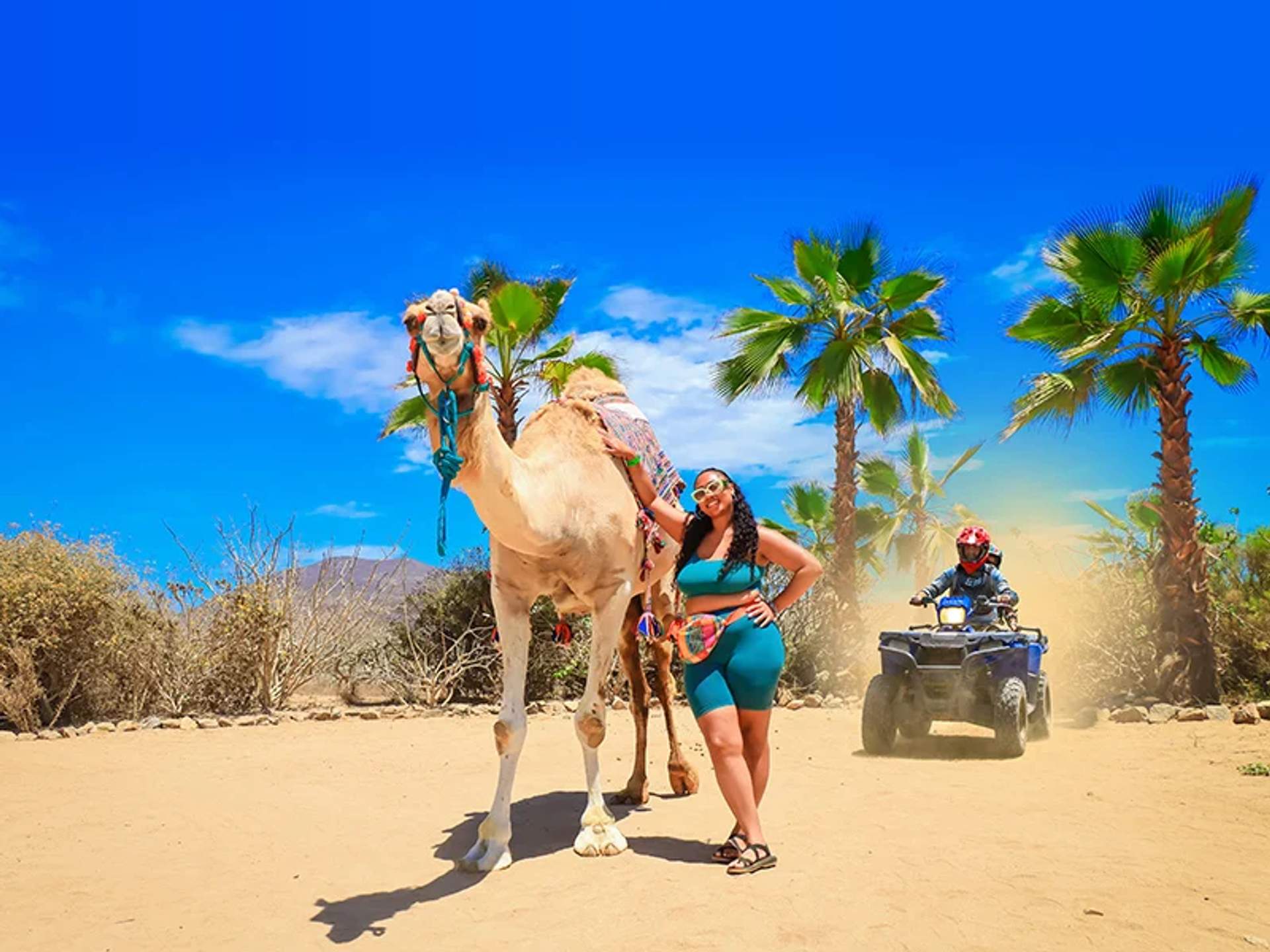 A woman poses with a camel under a clear blue sky, while a person rides an ATV in the background, surrounded by palm trees and desert scenery.