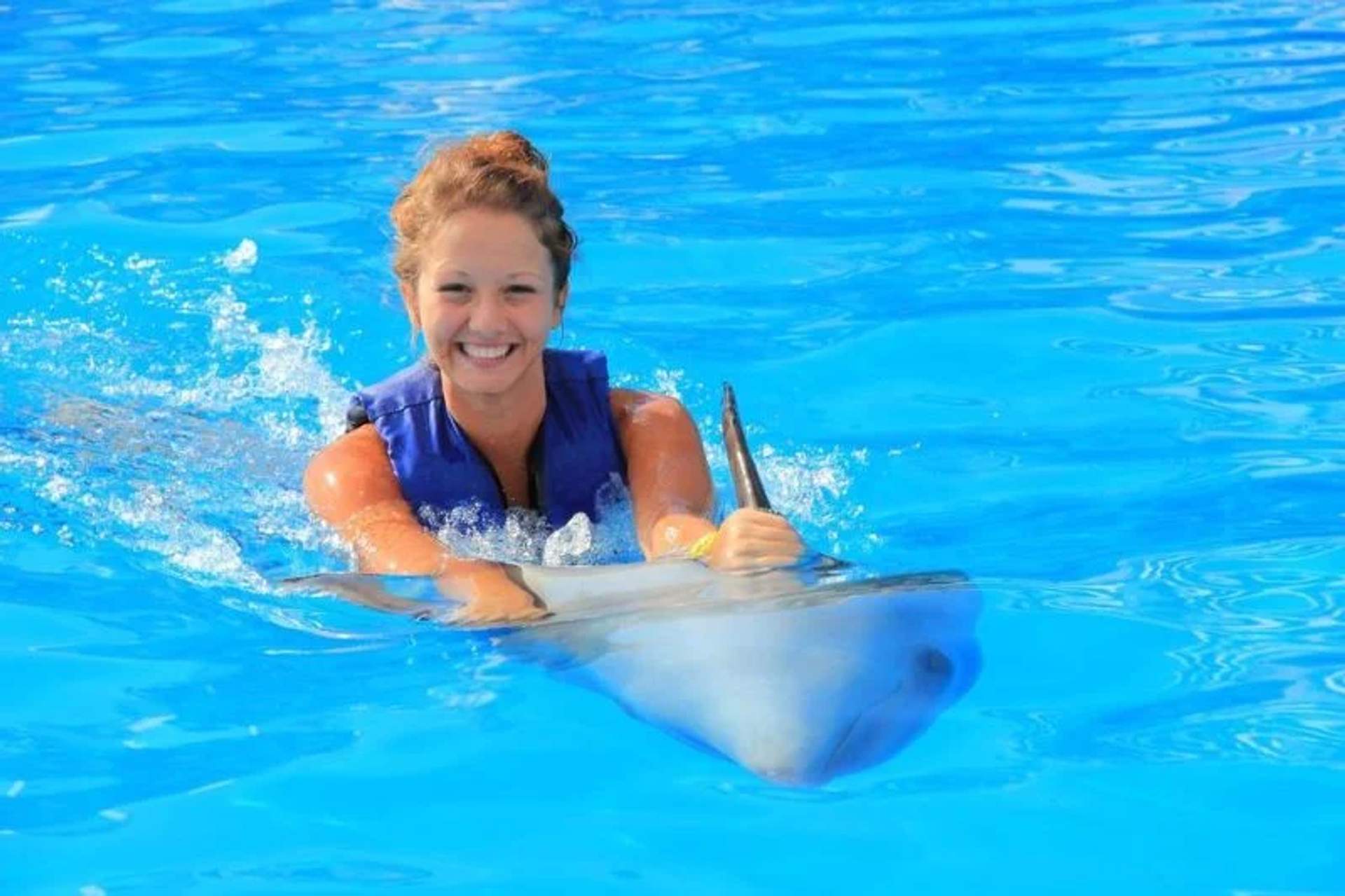A smiling woman in a blue life vest holds onto a dolphin while swimming in a bright blue pool. The dolphin's dorsal fin is visible, and both the woman and the dolphin appear to be enjoying the interaction.