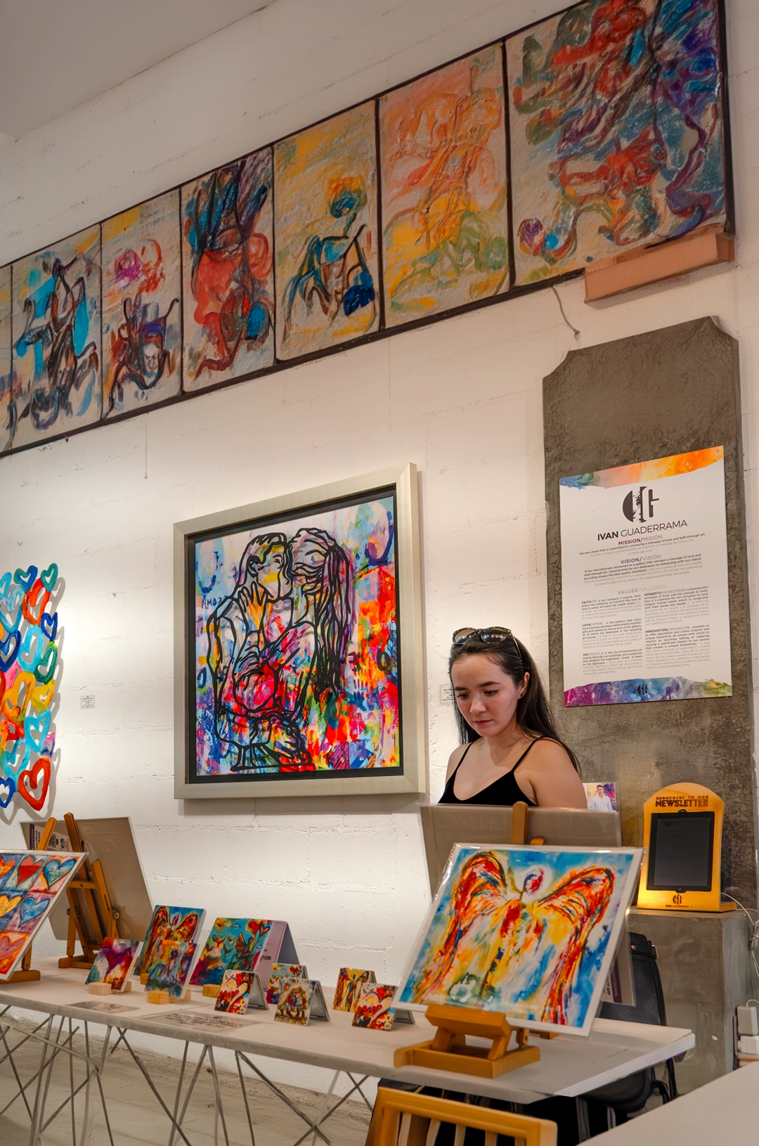 A woman browses colorful artwork in an art gallery, with vibrant paintings displayed on the walls and tables around her.