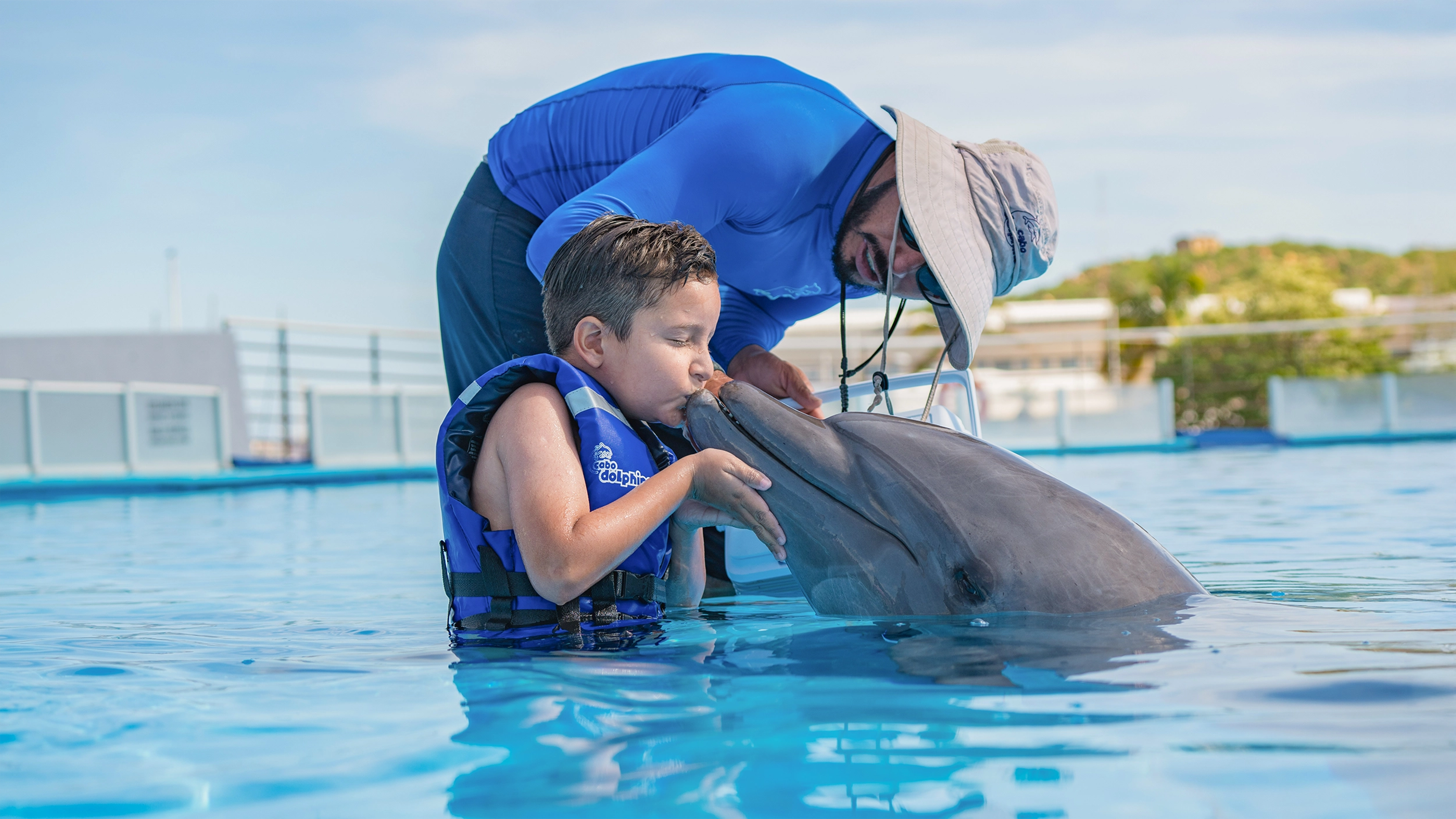 A young boy shares a sweet moment, kissing a dolphin during an interactive experience with a trainer.