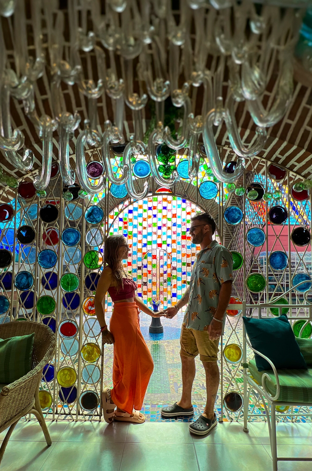 A couple holds hands in front of a colorful stained-glass archway, surrounded by brick walls and wicker chairs.