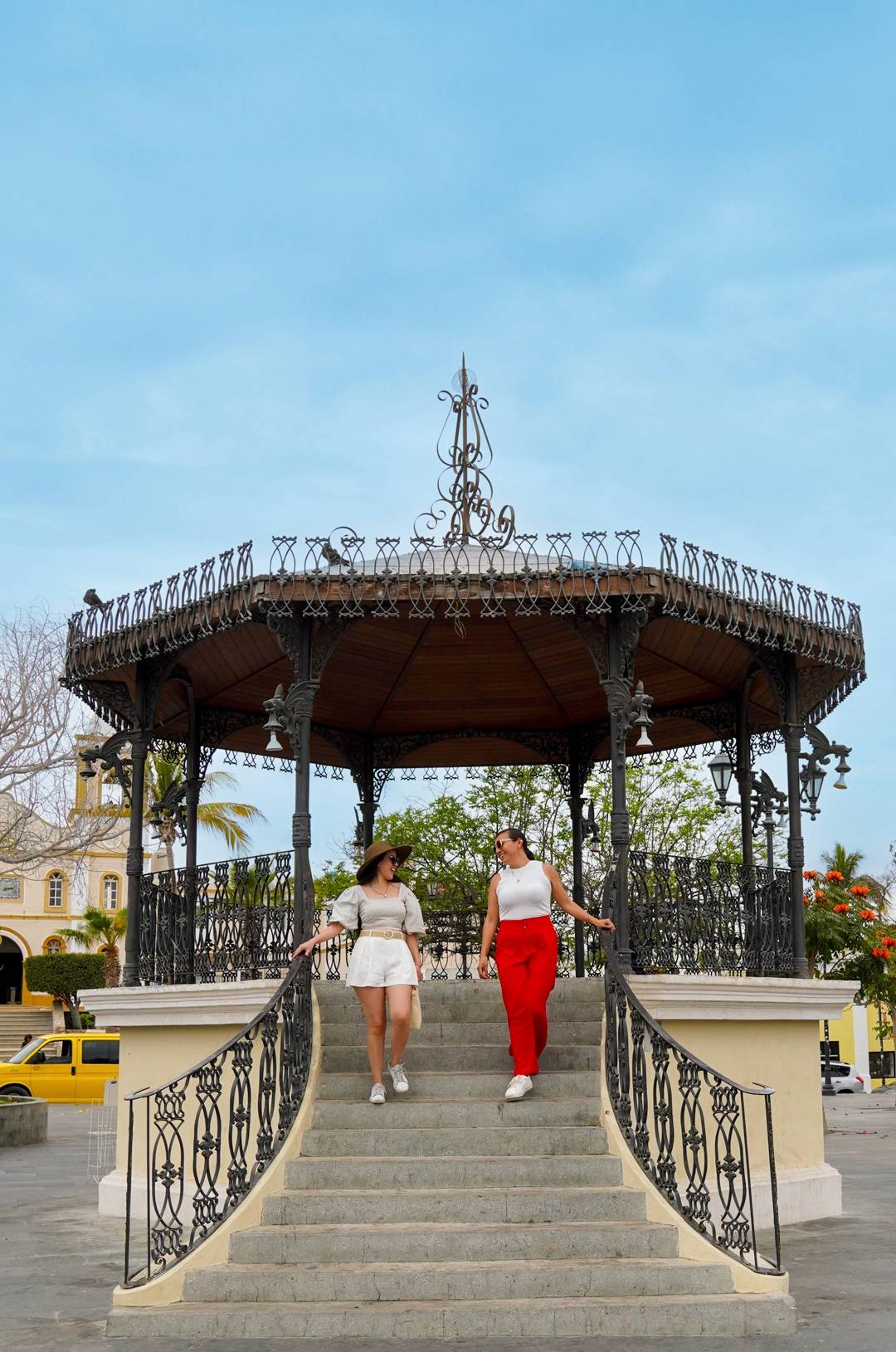 Two women descend the steps of a decorative gazebo with intricate ironwork in a sunny plaza.