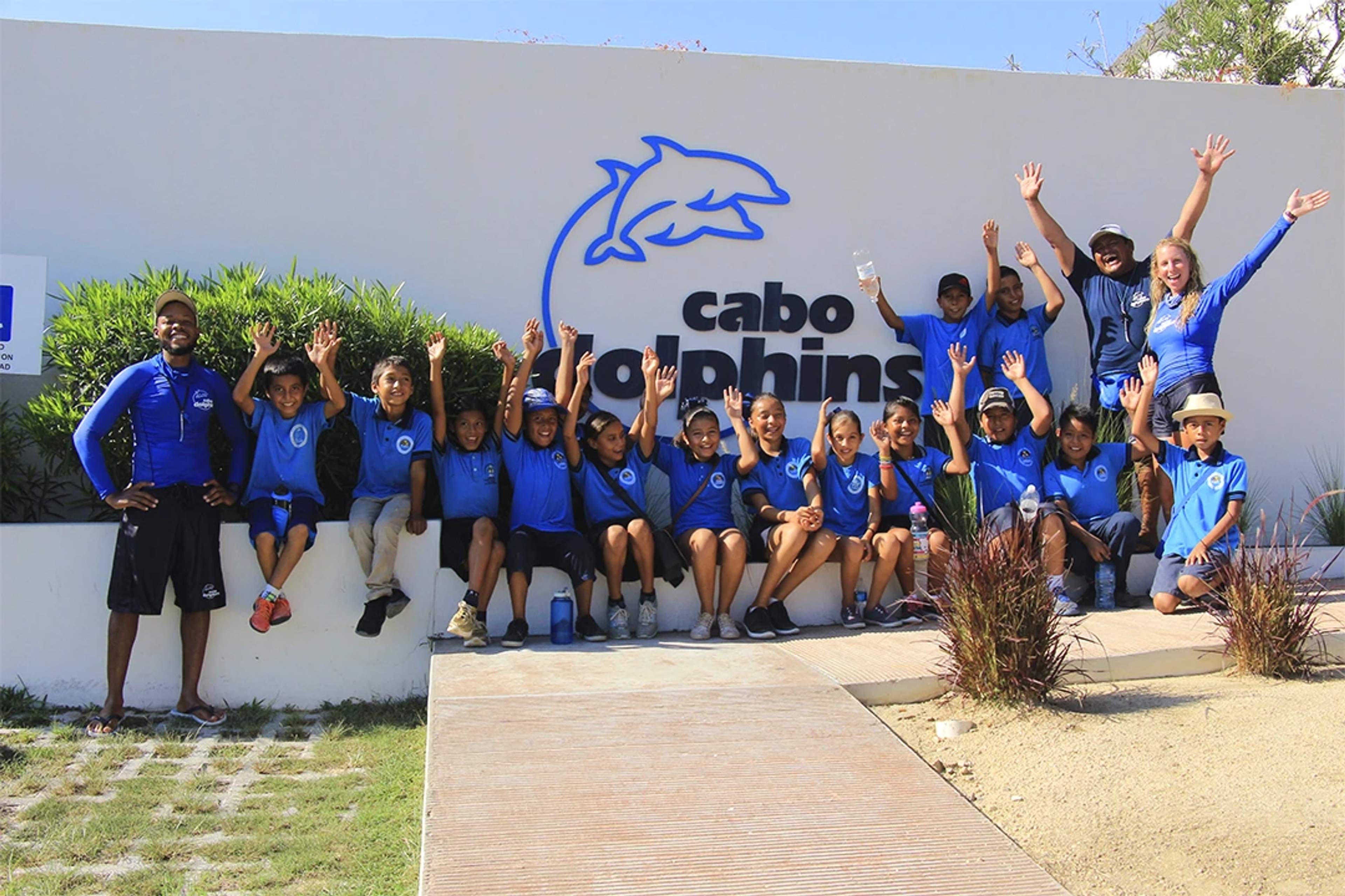 Group of smiling children and guides in blue uniforms raise their hands in front of the Cabo Dolphins logo.