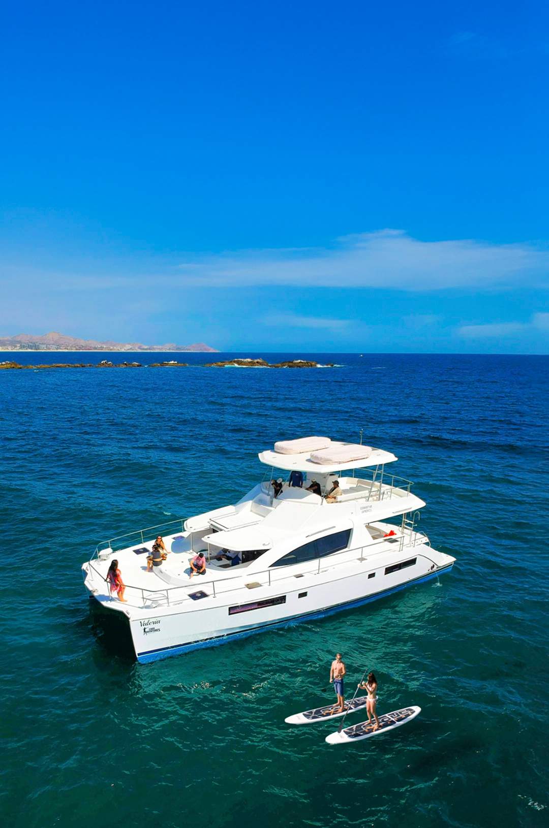 Luxury yacht with guests relaxing on deck, two paddleboarders nearby, all under a clear, sunny sky.