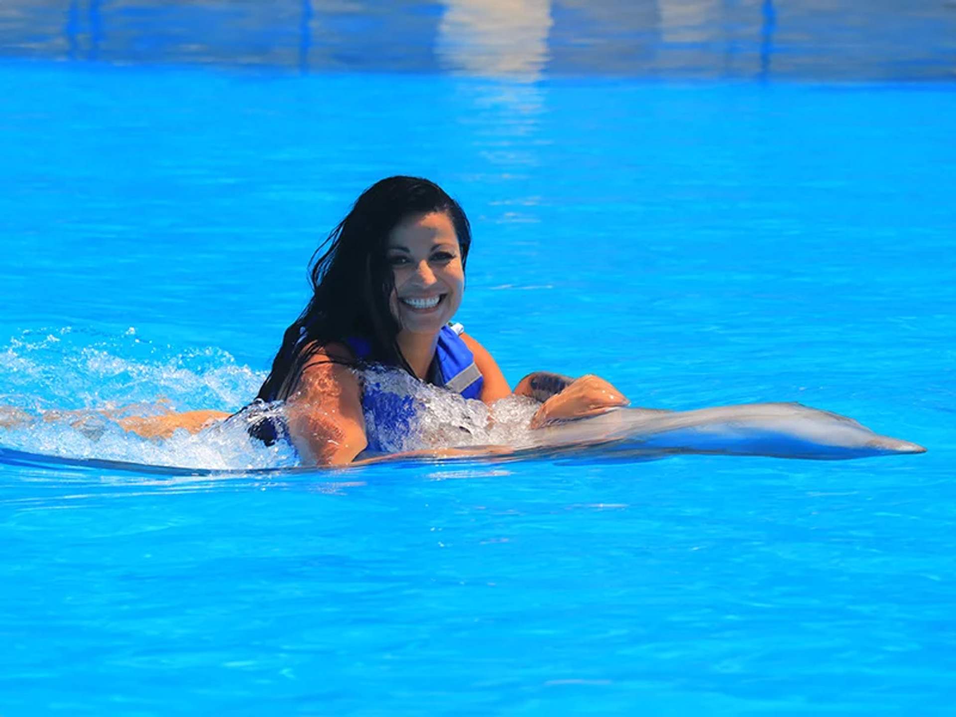 A woman with long dark hair smiles while swimming with a dolphin in a bright blue pool.