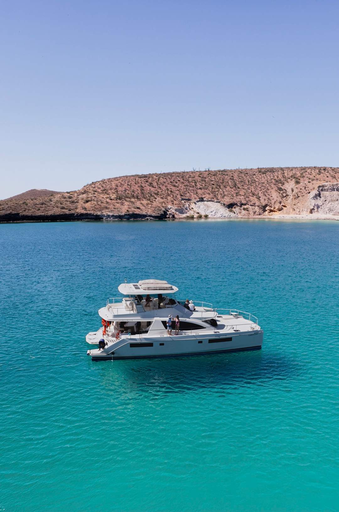 A white catamaran sailing on clear turquoise waters near a rocky coastline with a few people on board.