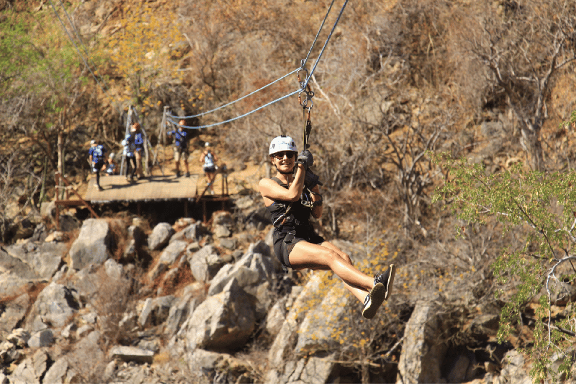 A person ziplining through a rocky and dry landscape, wearing a helmet and harness, with other people waiting on a platform in the background.