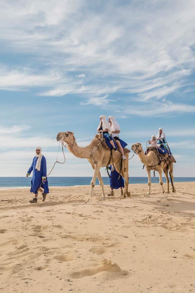 Man riding a camel led by a local guide in a desert landscape with cacti