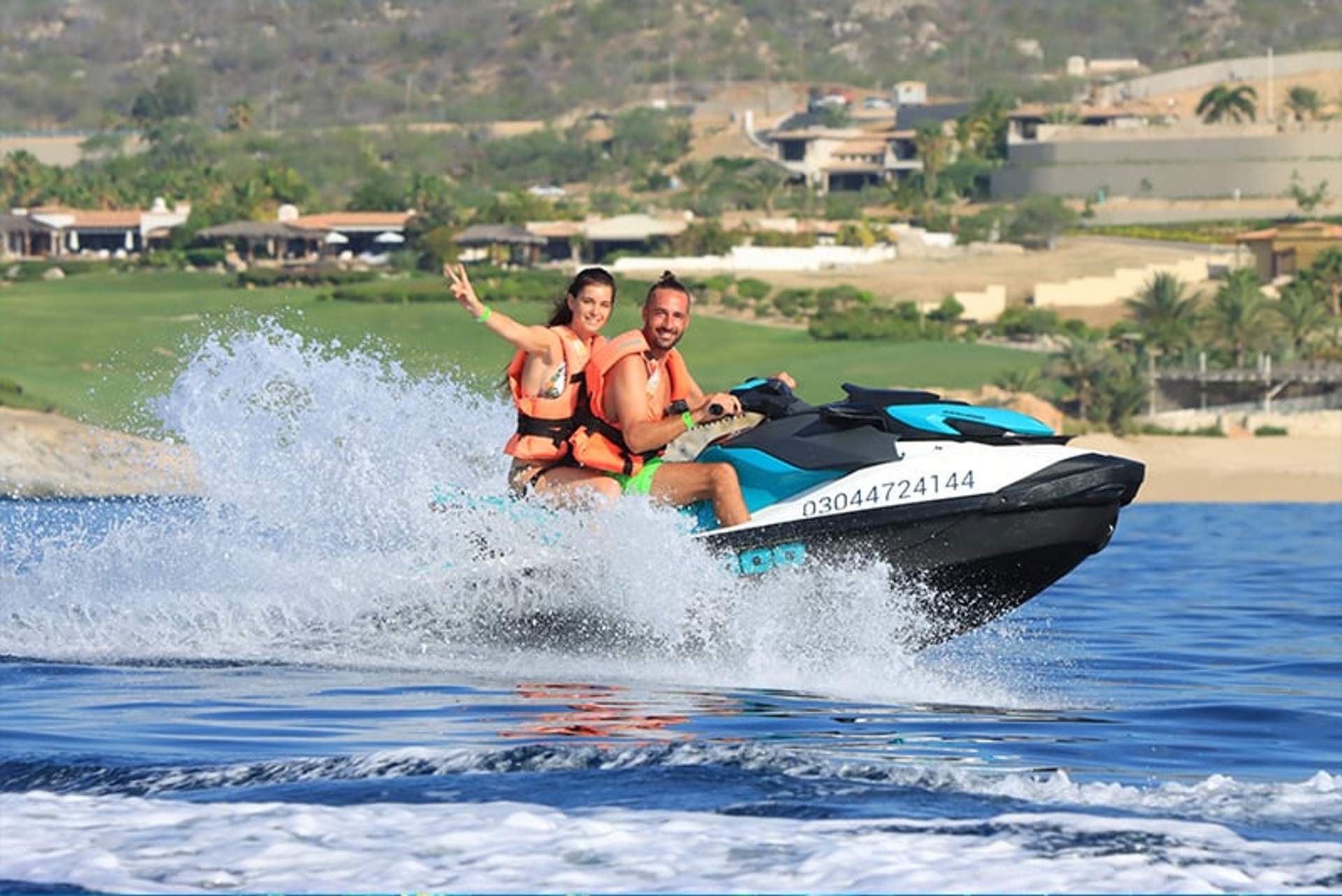 A smiling couple rides a jet ski on a sunny day, splashing through the water. They are both wearing orange life jackets, with scenic waterfront homes and green hills visible in the background.