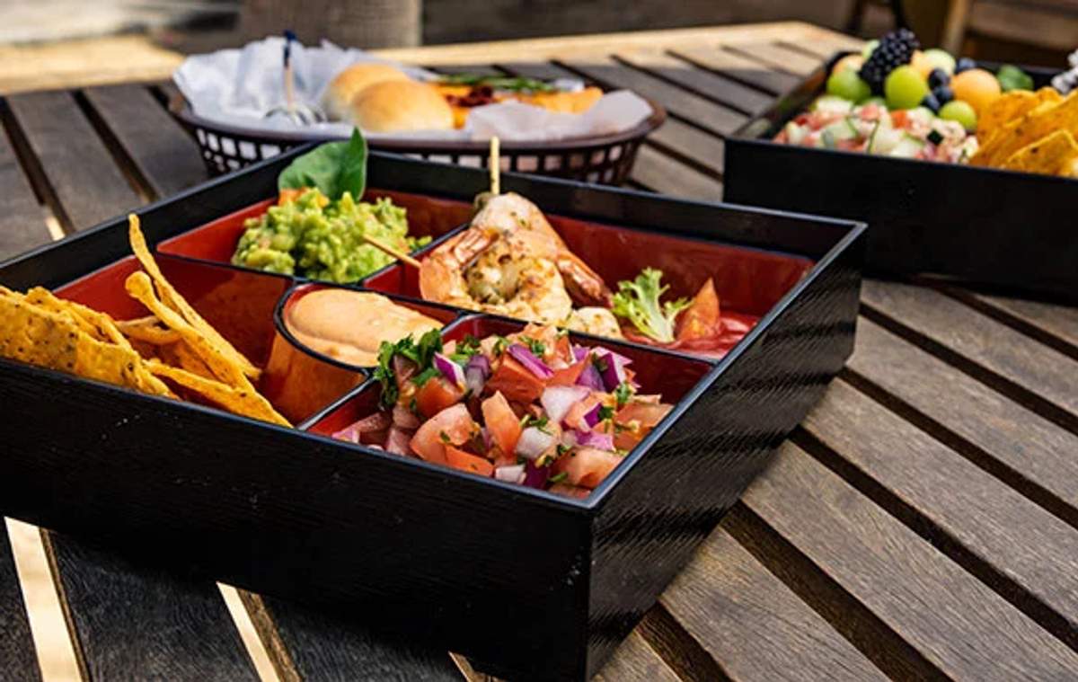 A bento box filled with a variety of fresh appetizers, including guacamole, shrimp, salsa, and chips, sits on a wooden table. The colorful and appetizing meal suggests an outdoor dining experience, possibly on a yacht or at a seaside location.