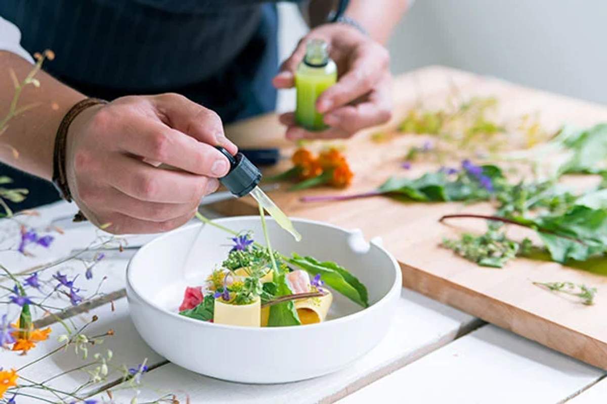 A chef carefully adding dressing to an artisanal salad with a dropper, surrounded by fresh herbs and flowers on a white table.