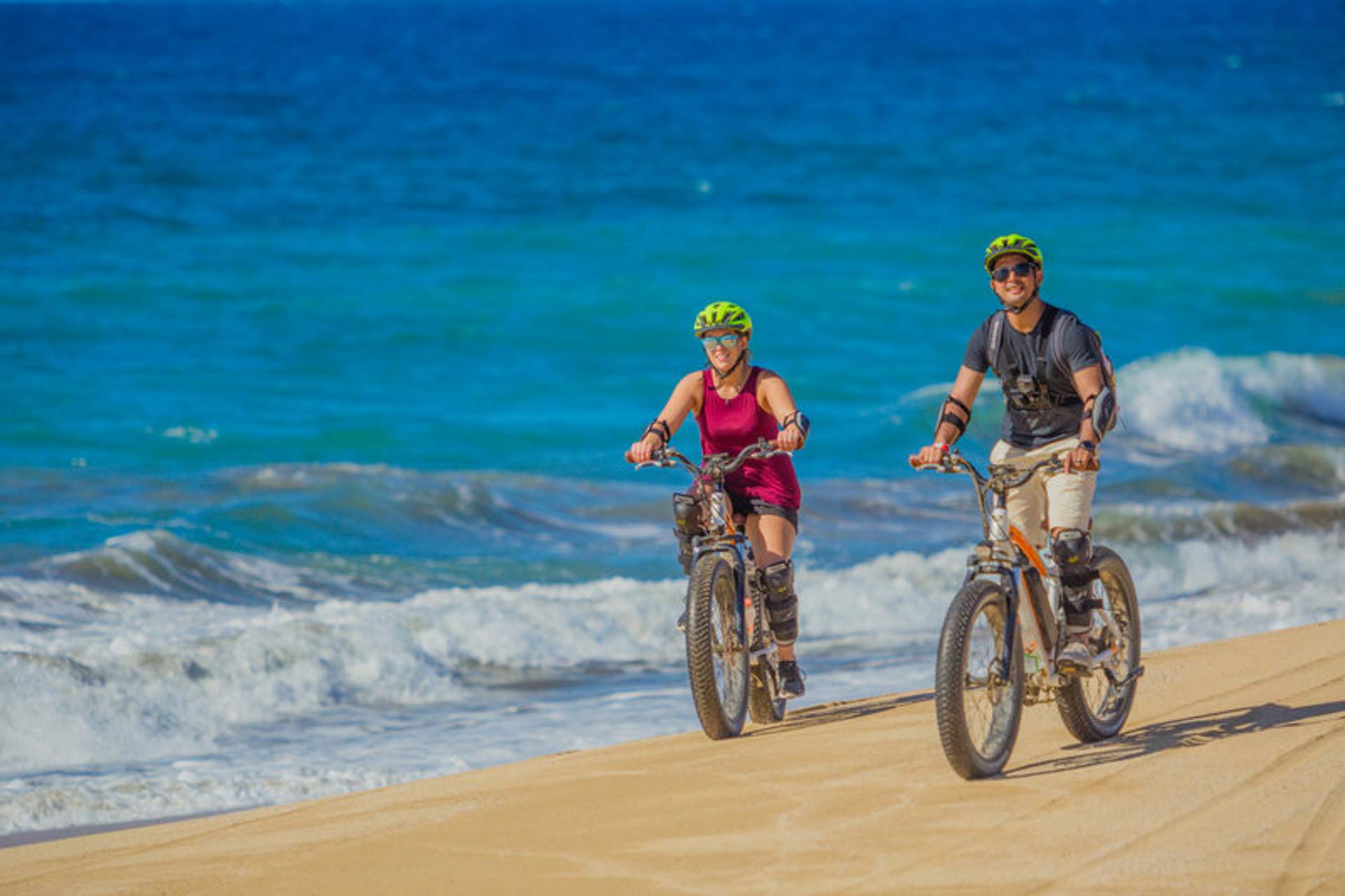 A man and a woman riding fat-tire bicycles along a sandy beach with vibrant blue ocean waves in the background. Both cyclists are wearing helmets and protective gear, enjoying a sunny day by the sea.
